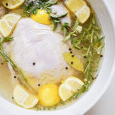 Raw chicken breast marinating in a bowl with lemon slices, fresh herbs (thyme, rosemary, bay leaves), black peppercorns, and a clear liquid.