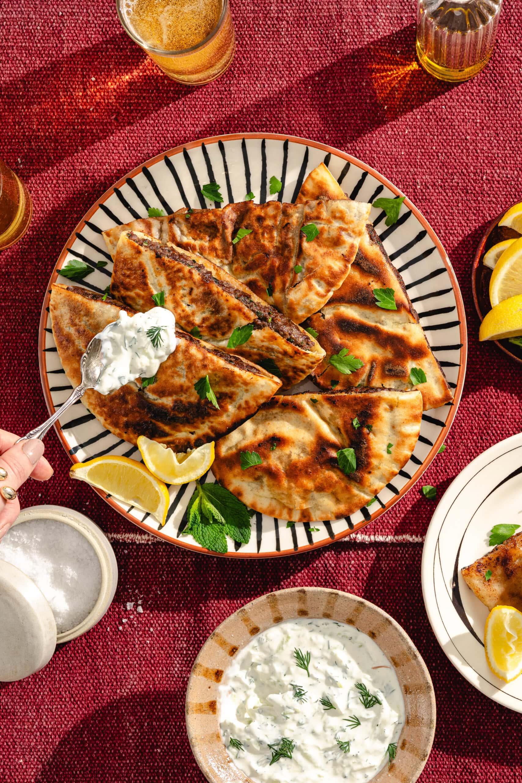 A plate of grilled flatbread wedges garnished with fresh herbs and lemon slices on a red tablecloth, with a hand serving tzatziki sauce. Surrounding the plate are drinks, extra lemon, and a bowl of tzatziki.