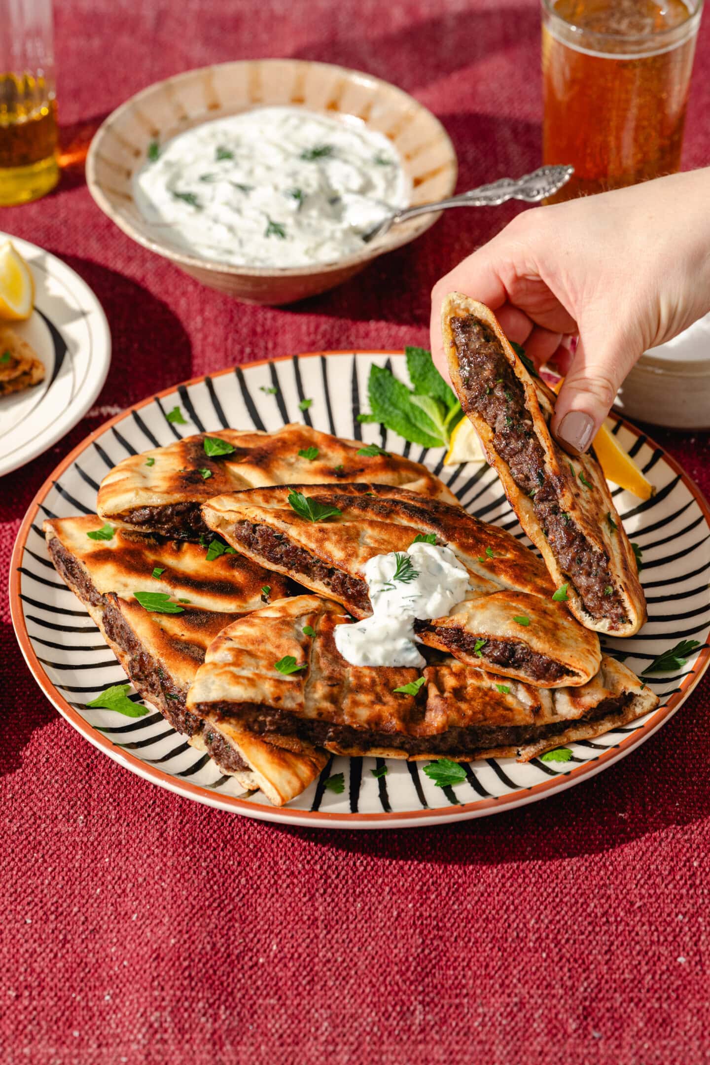 A hand picks up a piece of stuffed flatbread from a plate with several slices, topped with yogurt sauce and parsley. A bowl of yogurt dip, a lemon wedge, and a drink are on a red tablecloth nearby.