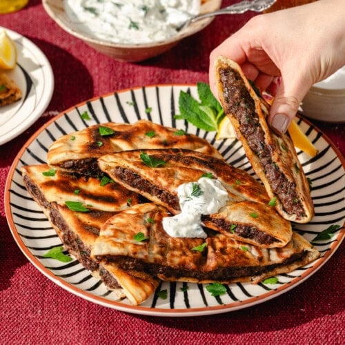 A hand picks up a piece of stuffed flatbread from a plate with several slices, topped with yogurt sauce and parsley. A bowl of yogurt dip, a lemon wedge, and a drink are on a red tablecloth nearby.
