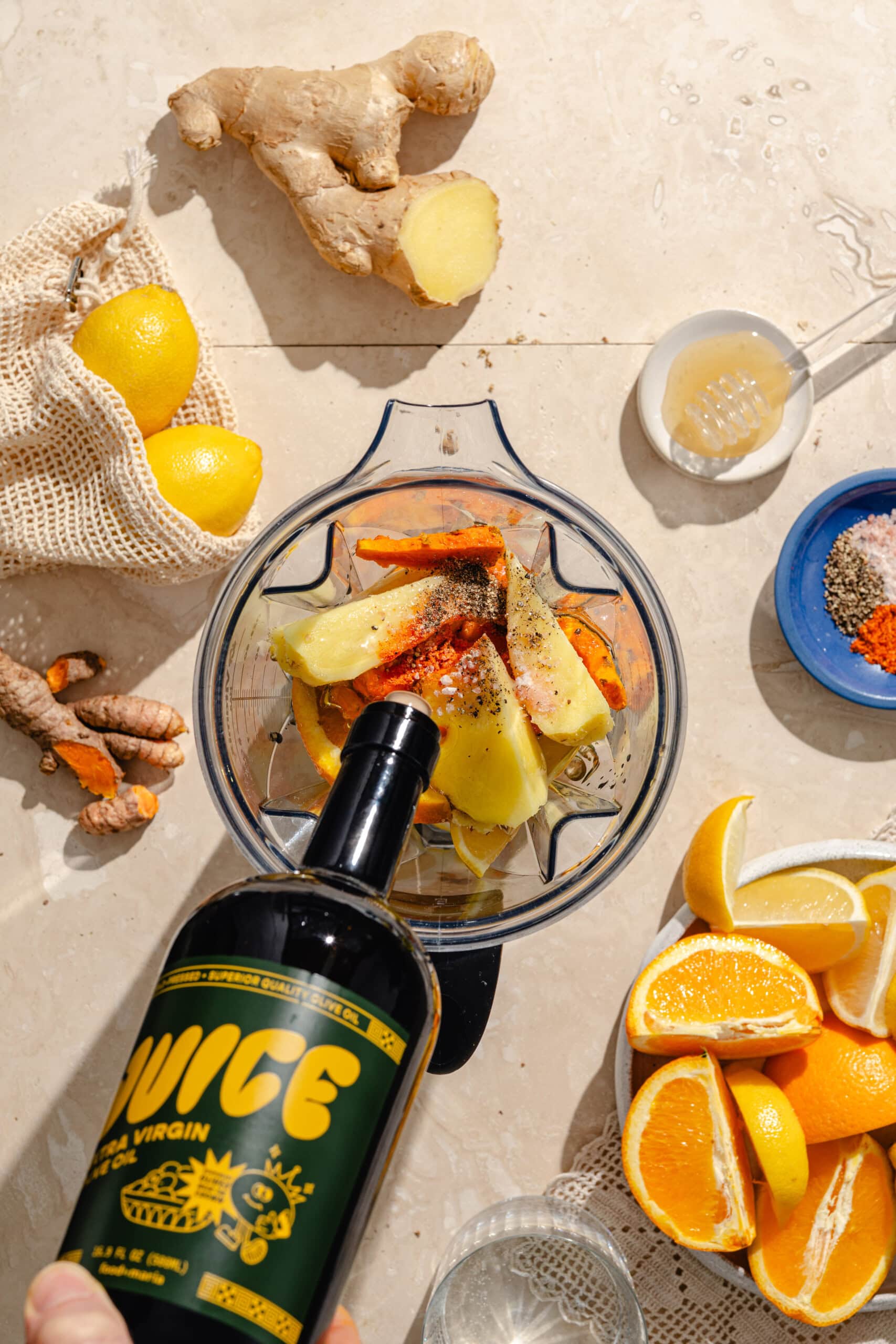 A hand pours olive oil from a bottle labeled JUICE into a blender filled with ginger, turmeric, apple, and spices. Surrounding are fresh oranges, ginger root, lemons, honey, salt, and a glass of water on a beige surface.