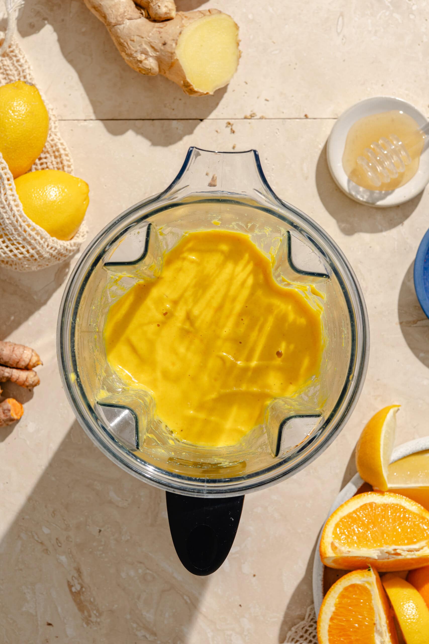 A blender filled with bright yellow smoothie sits on a light countertop, surrounded by fresh ginger, lemons, honey, turmeric, and orange slices, with sunlight casting shadows across the scene.