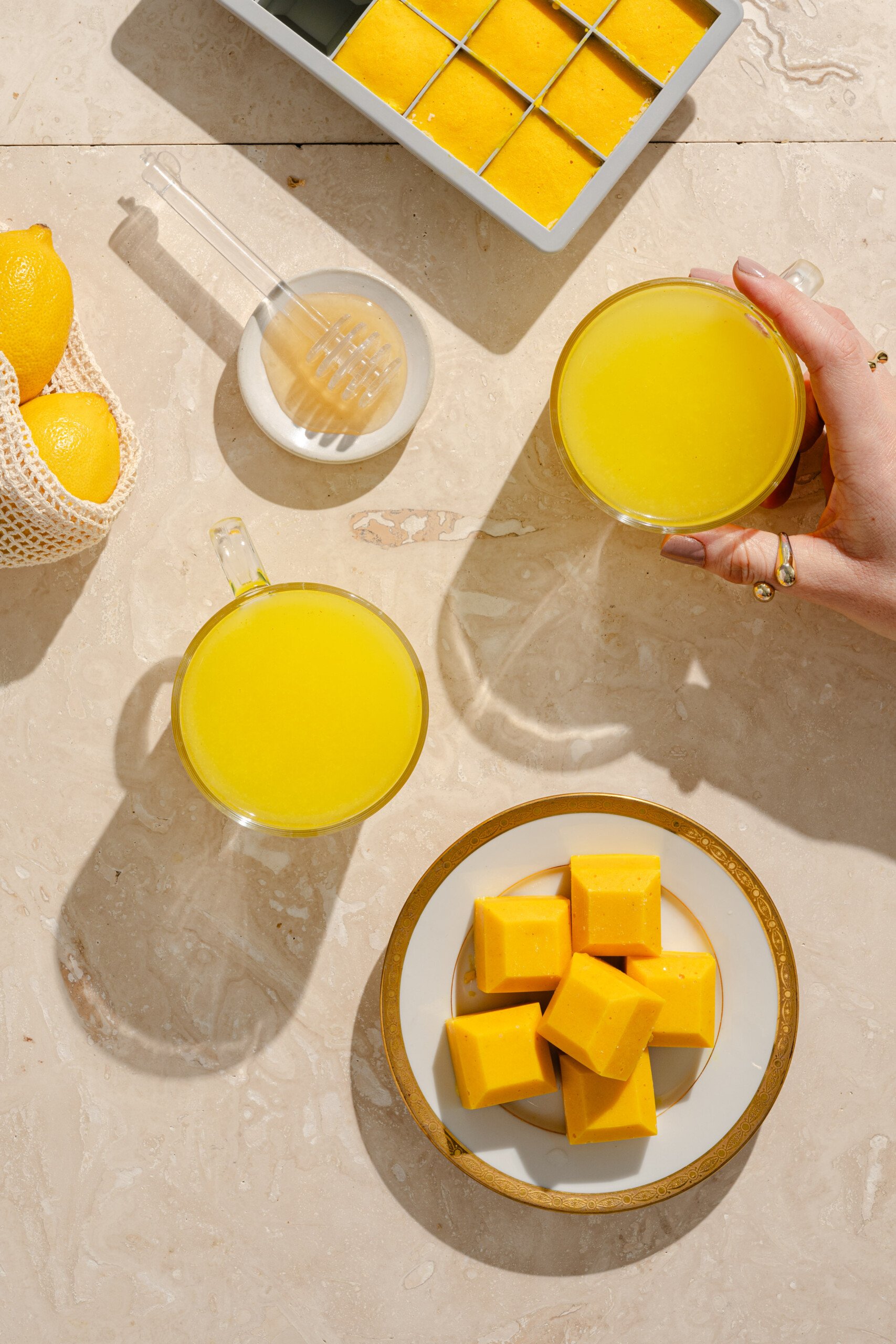Two mugs of yellow juice and a plate of yellow ice cubes on a marble surface, with a hand holding one mug, lemons in a bag, a honey dipper, and an ice cube tray also visible. Bright natural lighting.