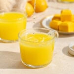 A glass cup filled with yellow juice sits on a light surface, with juice droplets falling into it. Behind it are another glass of juice, lemon cubes on a plate, and whole lemons in soft focus.