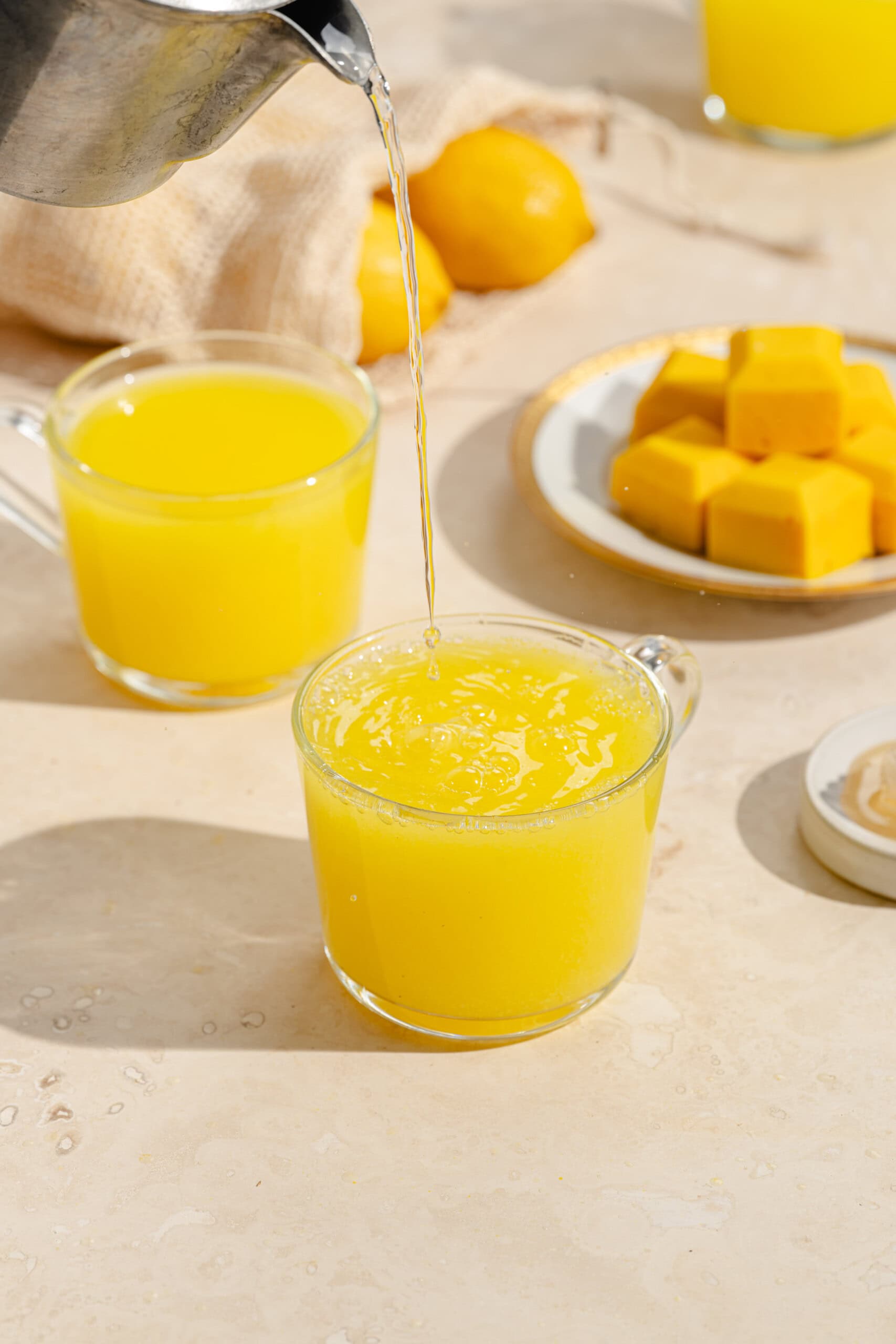 A clear glass mug is being filled with yellow juice. Another mug of juice, a plate with yellow cubes, lemons, and a beige cloth are on a light-colored surface in the background.
