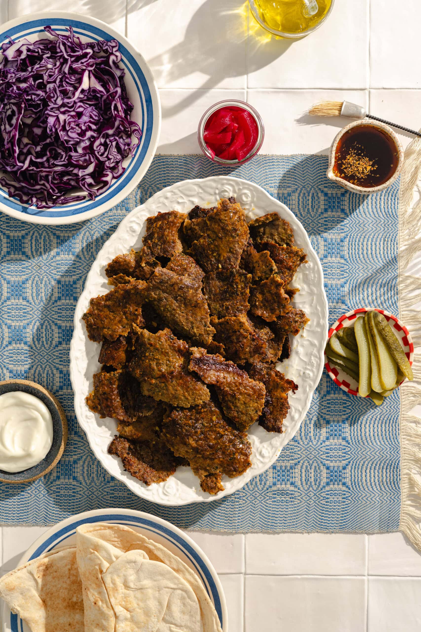 A table set with a platter of cooked meat, a bowl of shredded red cabbage, pickles, pita bread, a dish of sauce, sliced red peppers, and a bottle of olive oil on a blue woven placemat.