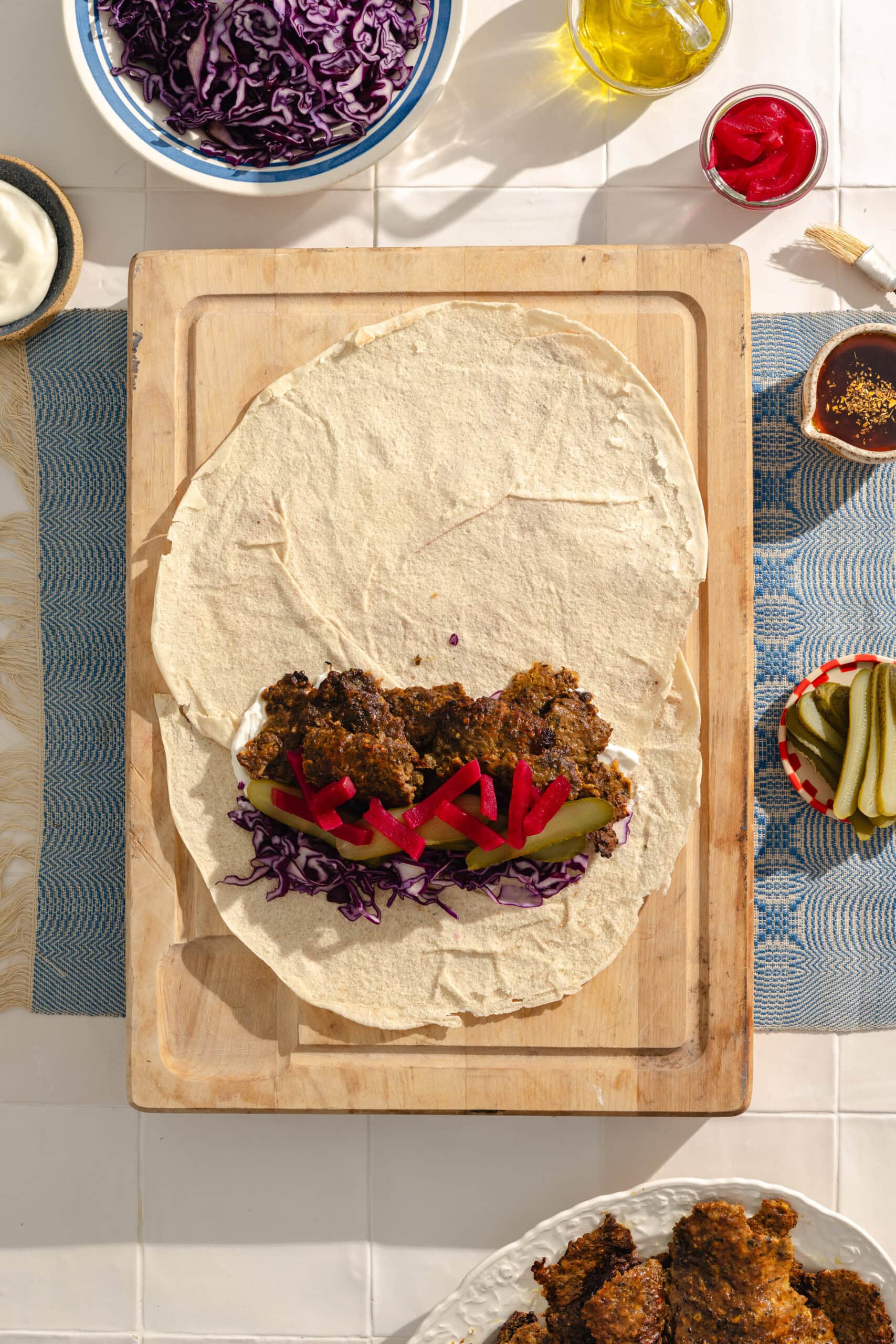 A tortilla on a wooden board is topped with breaded chicken, sliced pickles, shredded red cabbage, and red bell pepper strips. Surrounding dishes hold pickles, cabbage, oil, sauce, and more chicken.