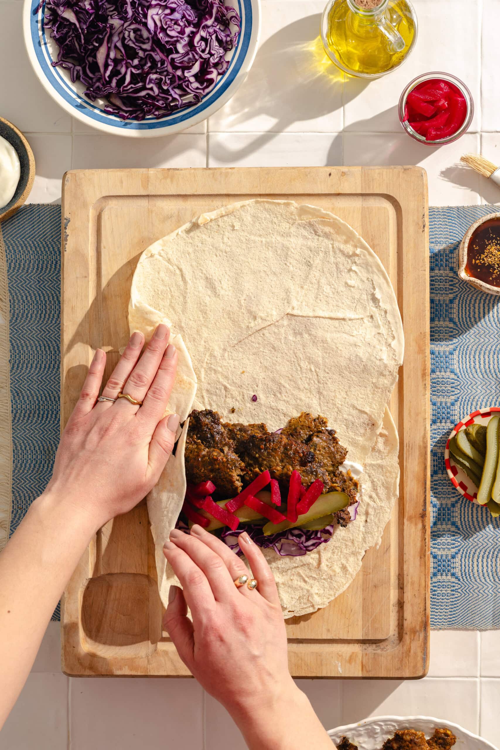 A person folds a large tortilla wrap filled with shredded red cabbage, sliced pickles, and a dark spiced meat mixture on a wooden board, surrounded by bowls of cabbage, oil, pickled vegetables, and condiments.