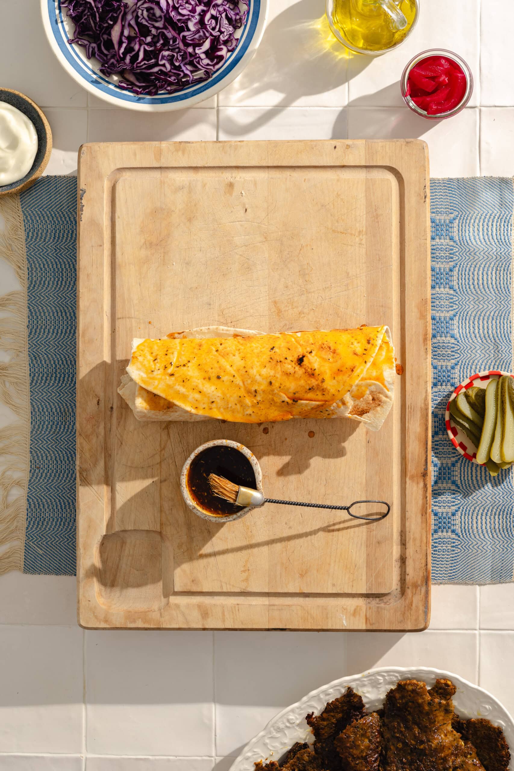 A large wrap topped with melted cheese sits on a wooden cutting board. Nearby are bowls of sliced pickles, shredded red cabbage, ketchup, oil, and a plate of fried food, all on a white tiled surface.