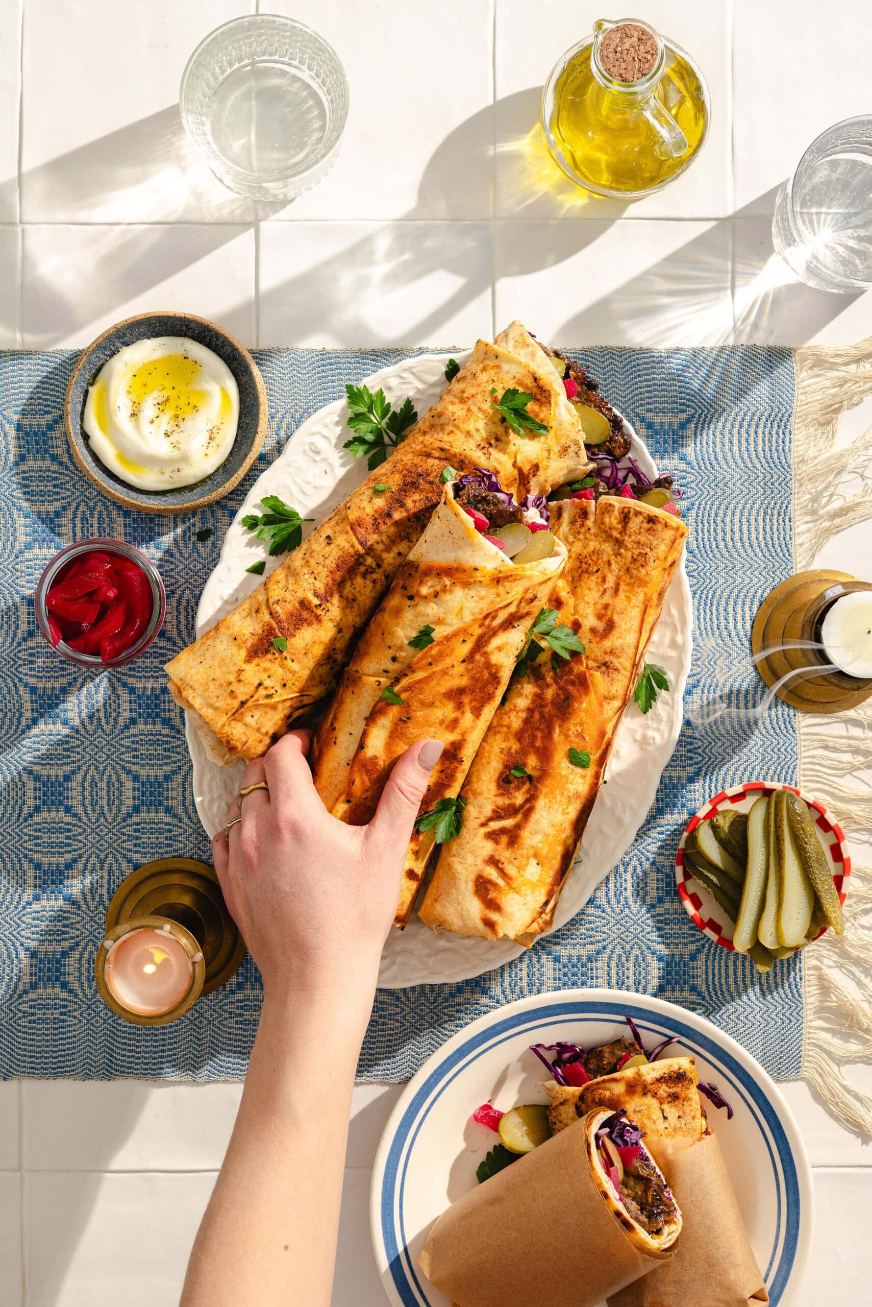 A hand reaches for a toasted wrap filled with colorful vegetables on a white platter, surrounded by small bowls of pickles, yogurt, and drinks on a bright, sunlit table.