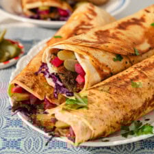 Close-up of two large wraps filled with seasoned meat, sliced pickles, red cabbage, and herbs, served on a white plate with a blue patterned tablecloth and garnished with fresh parsley.