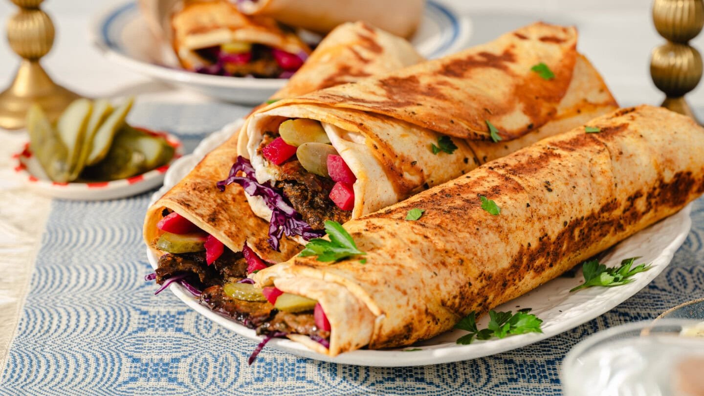 Three large shawarma wraps filled with sliced meat, pickles, red cabbage, and vegetables are arranged on a white plate, garnished with parsley, on a patterned blue tablecloth.