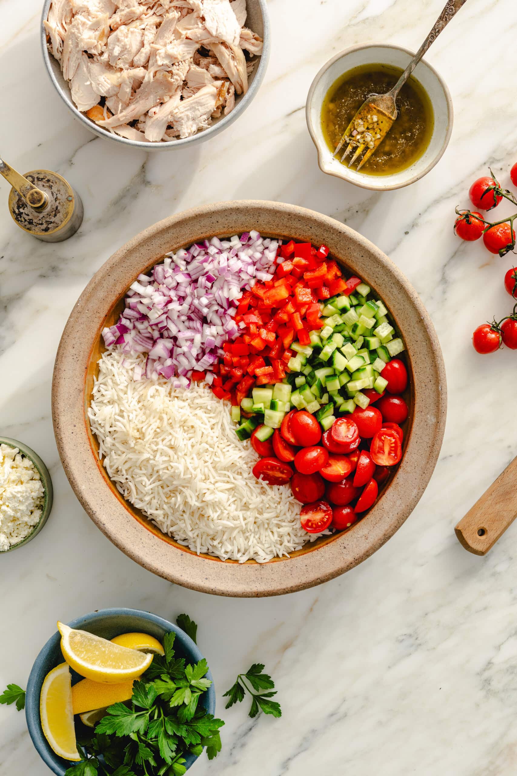 A bowl filled with rice, chopped red onion, red bell pepper, cucumber, and halved cherry tomatoes sits on a marble counter, surrounded by bowls of lemon wedges, herbs, feta, shredded chicken, and a small bowl of dressing.