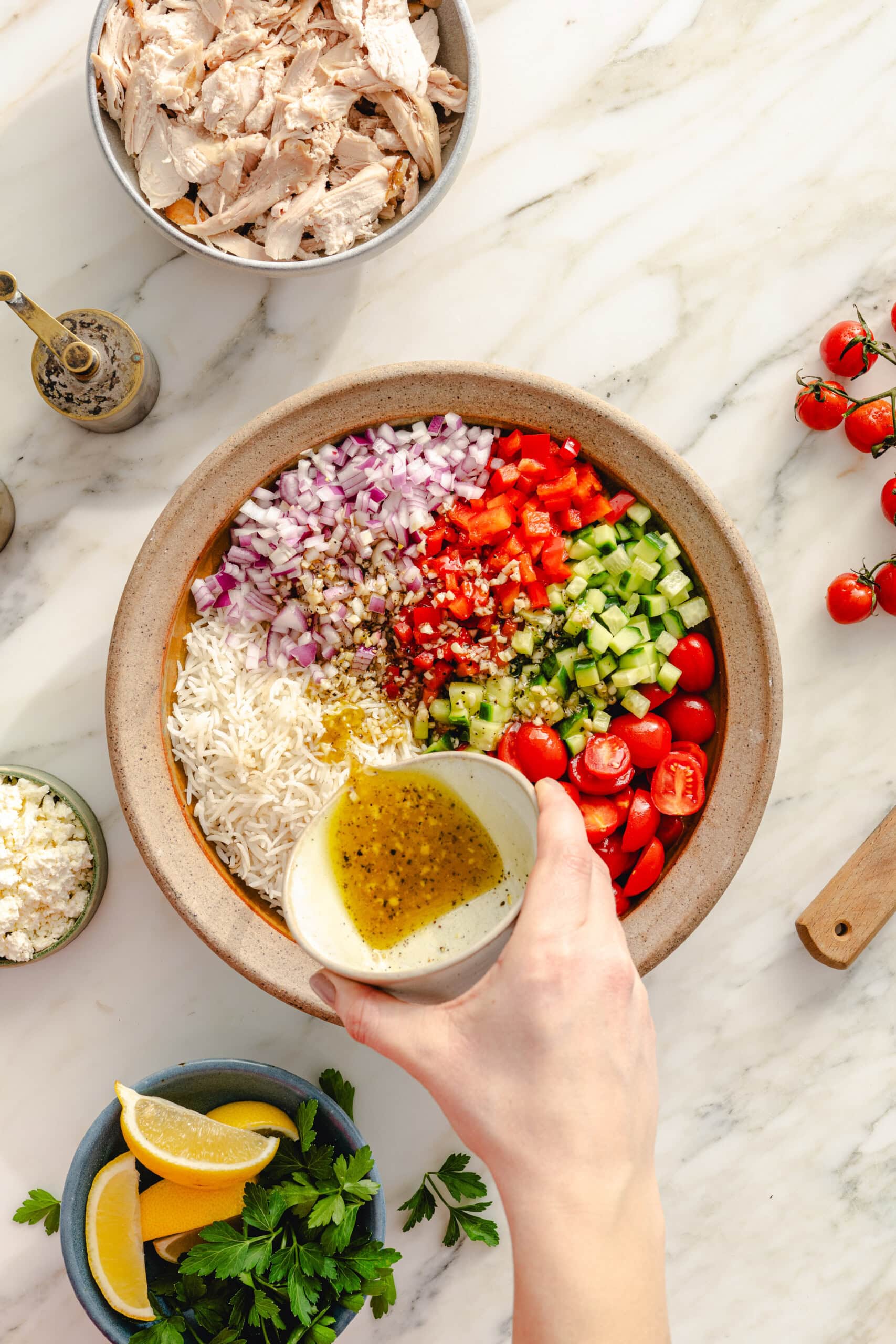 A hand pours vinaigrette over a bowl filled with chopped vegetables, rice, and herbs. Surrounding the bowl are ingredients like shredded chicken, feta cheese, lemon wedges, and fresh parsley on a marble countertop.