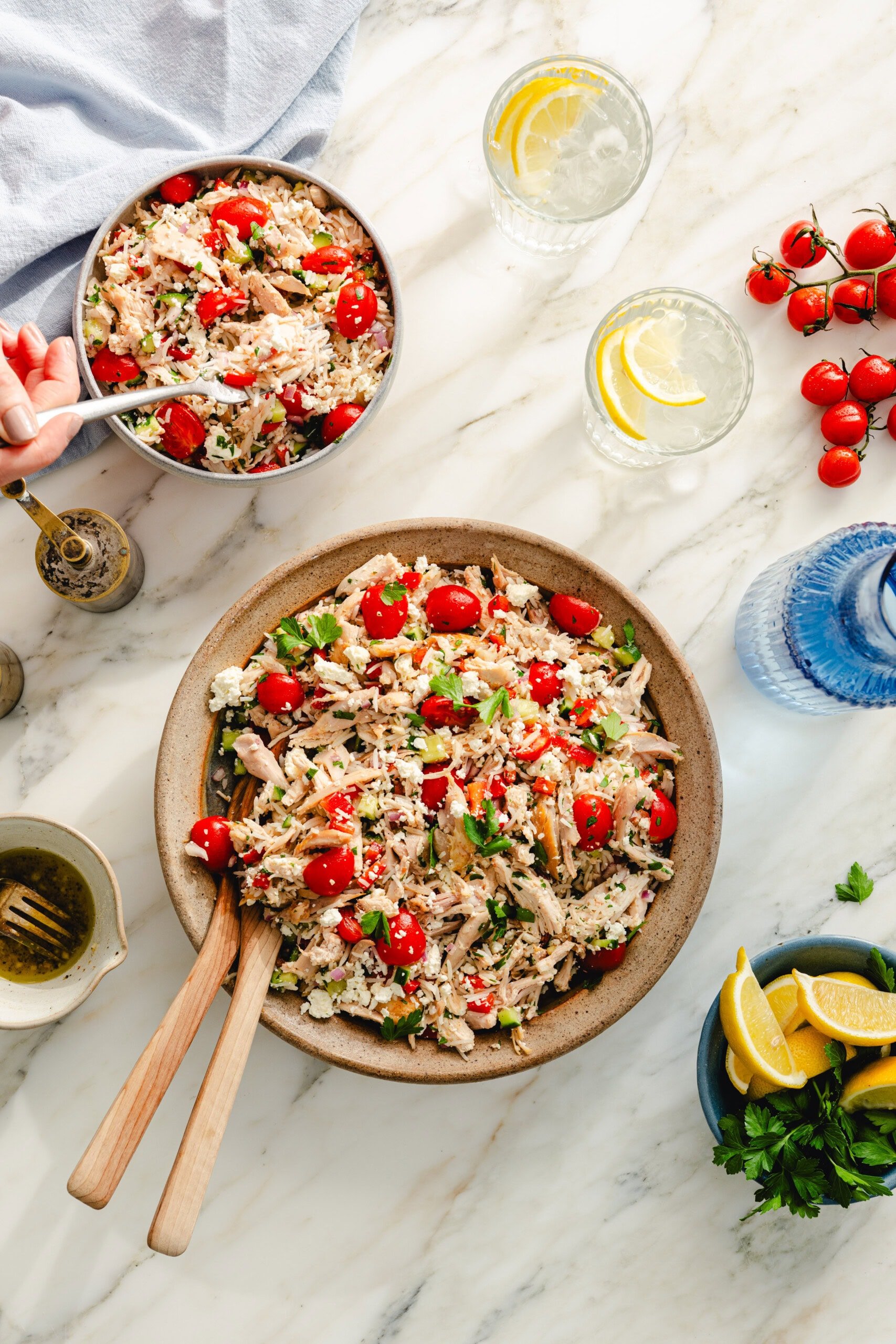 A large bowl of orzo salad with shredded chicken, cherry tomatoes, feta cheese, and herbs sits on a marble table. A hand serves a smaller bowl. Sliced lemons, cherry tomatoes, and glasses of water with lemon are nearby.