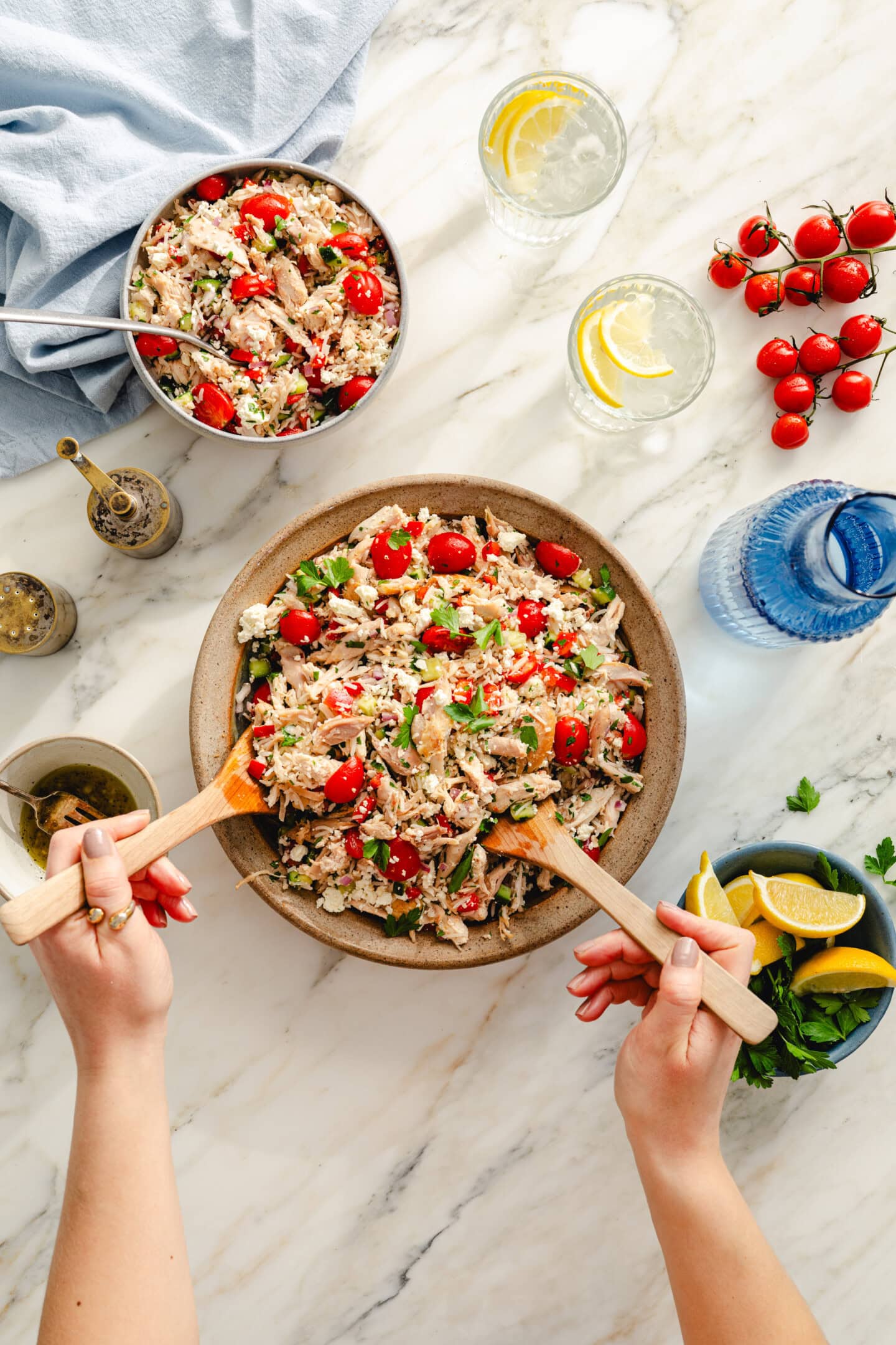 A person serves a fresh salad with tomatoes, greens, and shredded chicken from a large bowl, surrounded by ingredients, lemon slices, water, and smaller salad serving on a marble countertop.