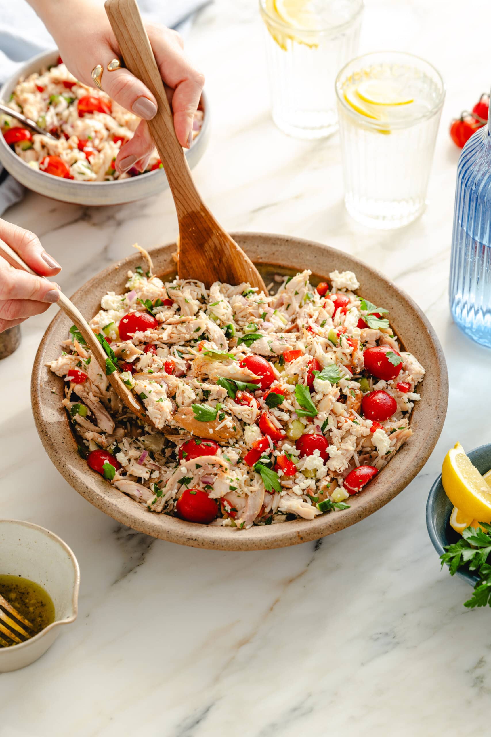 Hands toss a salad with shredded chicken, cherry tomatoes, rice, herbs, and feta in a large bowl with wooden utensils. A smaller bowl of salad, lemon wedges, and glasses of water with lemon are nearby on a marble surface.