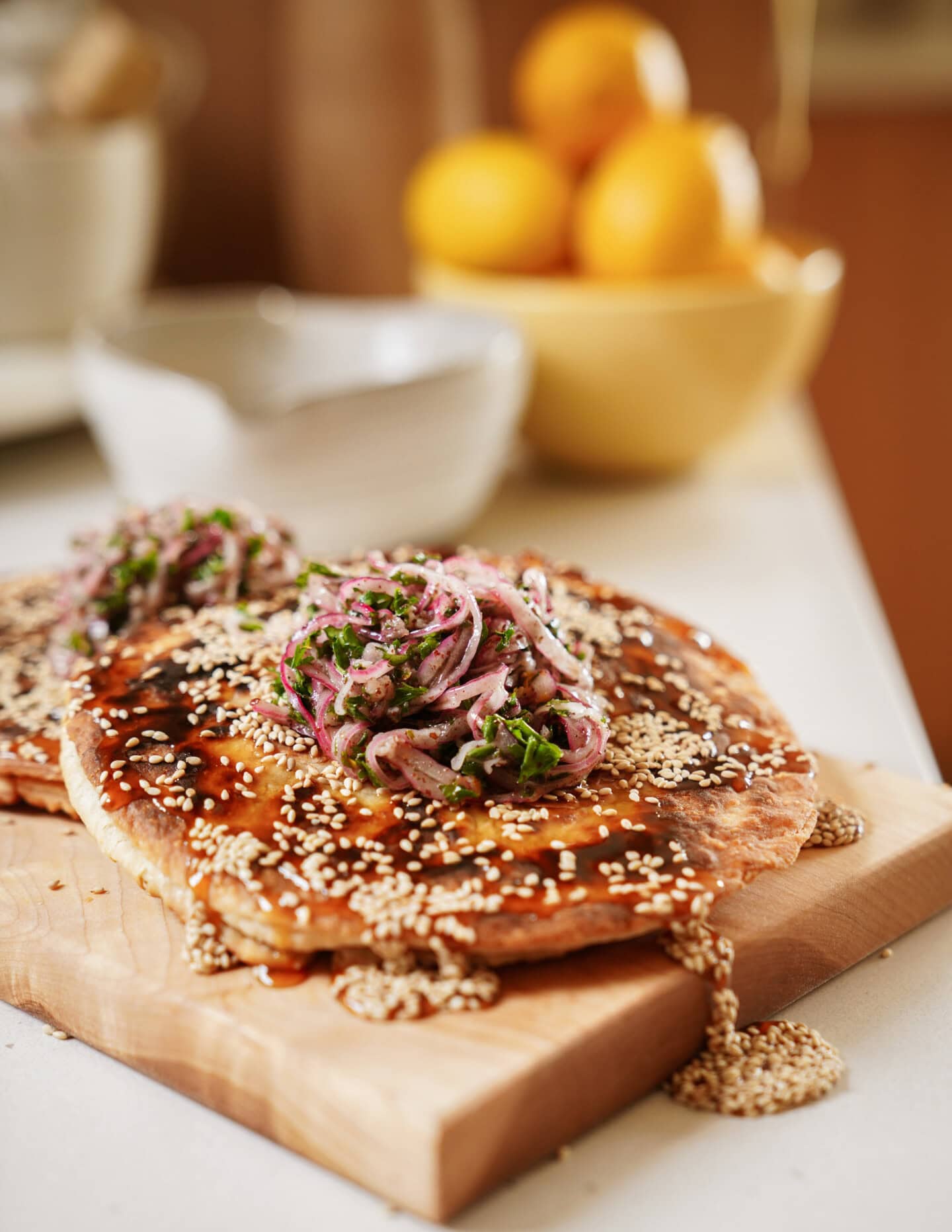 Flatbreads topped with sesame seeds, sauce, and a garnish of thinly sliced onions and herbs sit on a wooden board. In the background, there is a bowl of yellow lemons and a ceramic bowl on a kitchen counter.