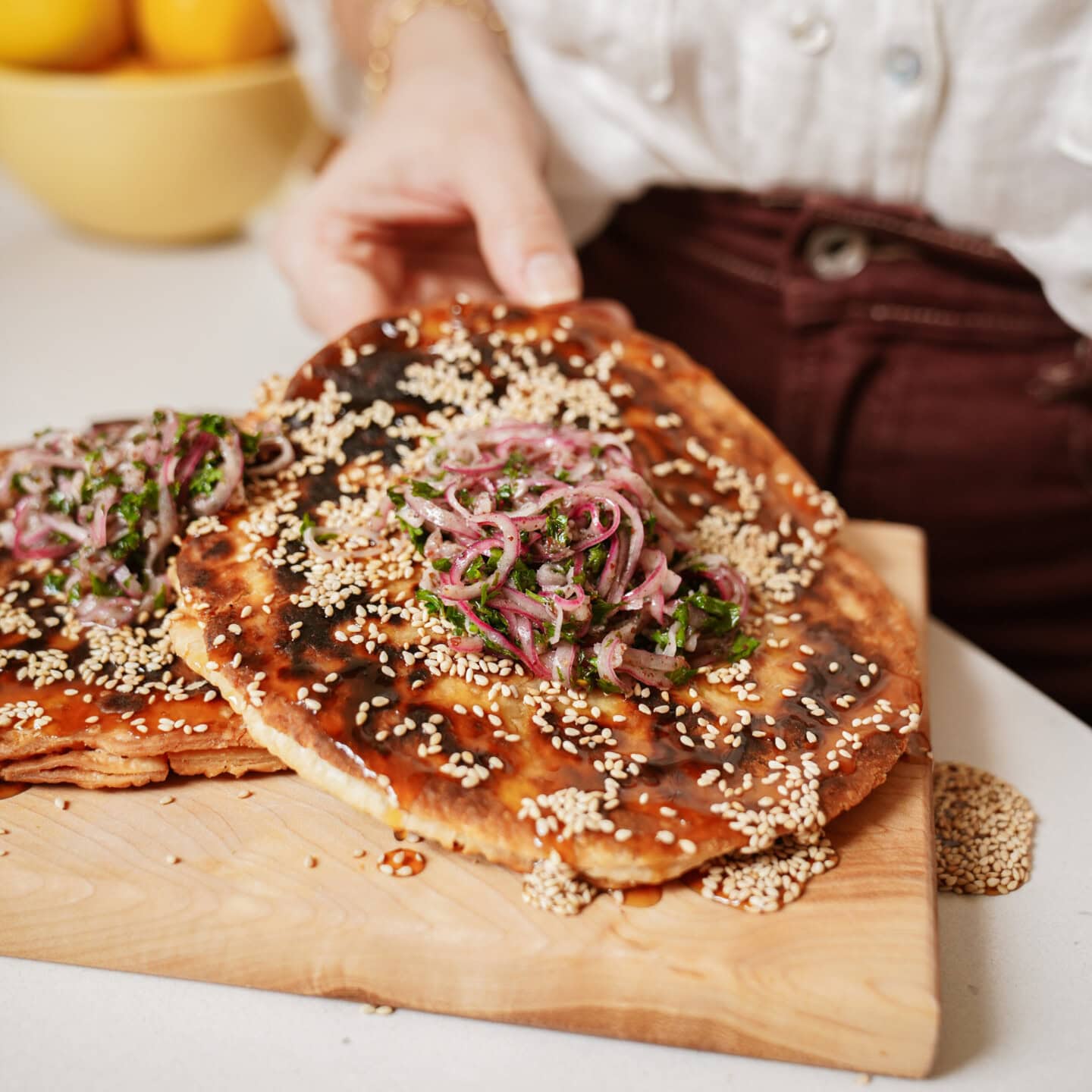 A person holding flatbread topped with sesame seeds, pickled red onions, and fresh herbs on a wooden cutting board. A bowl of lemons is blurred in the background.