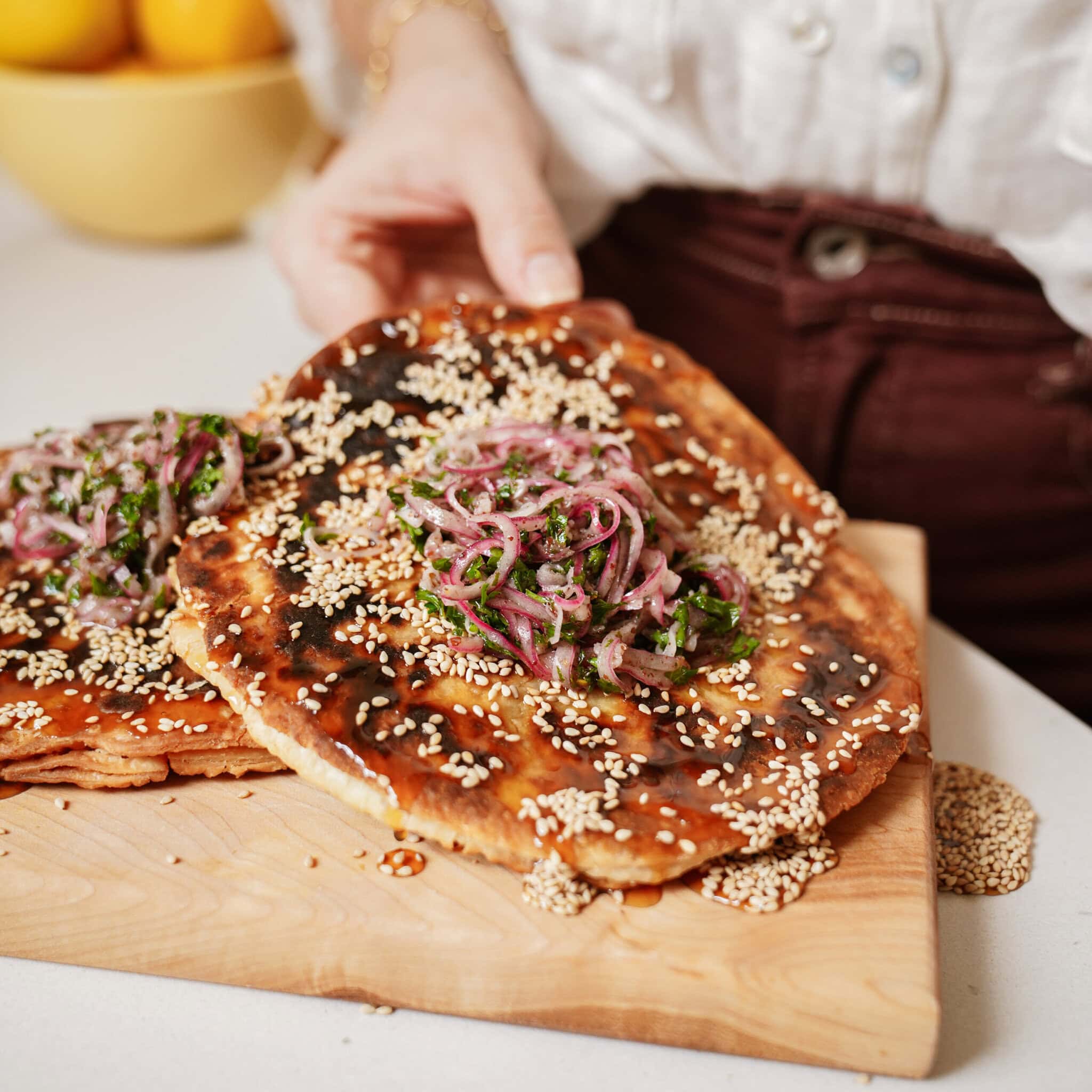 A person holding flatbread topped with sesame seeds, pickled red onions, and fresh herbs on a wooden cutting board. A bowl of lemons is blurred in the background.