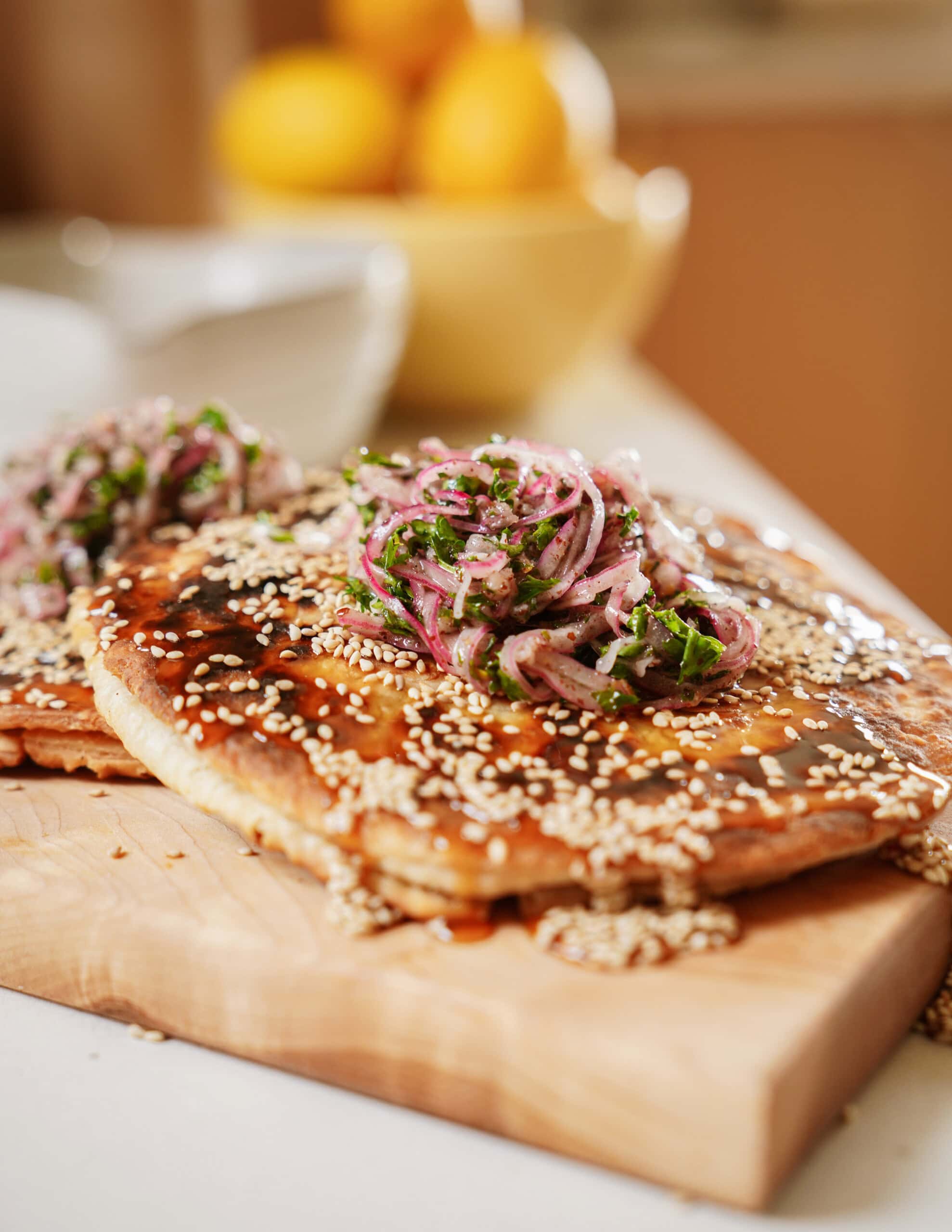 A close-up of flatbread topped with sesame seeds, red onions, and herbs, served on a wooden board with a blurred bowl of lemons in the background.