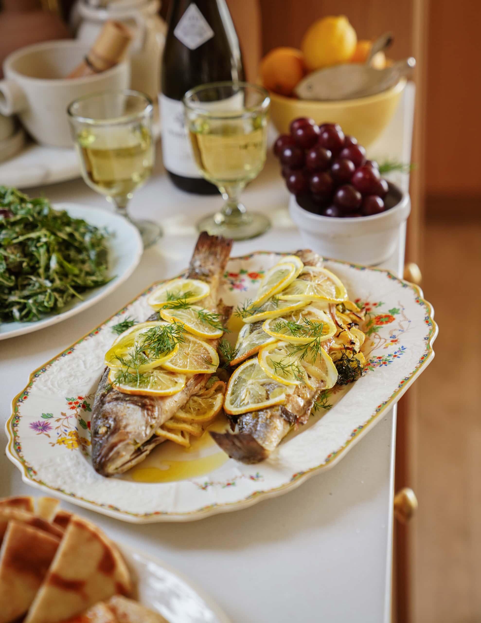 A platter with two whole roasted fish garnished with lemon slices and fresh dill, accompanied by glasses of white wine, a bowl of grapes, salad, and flatbread on a white table.