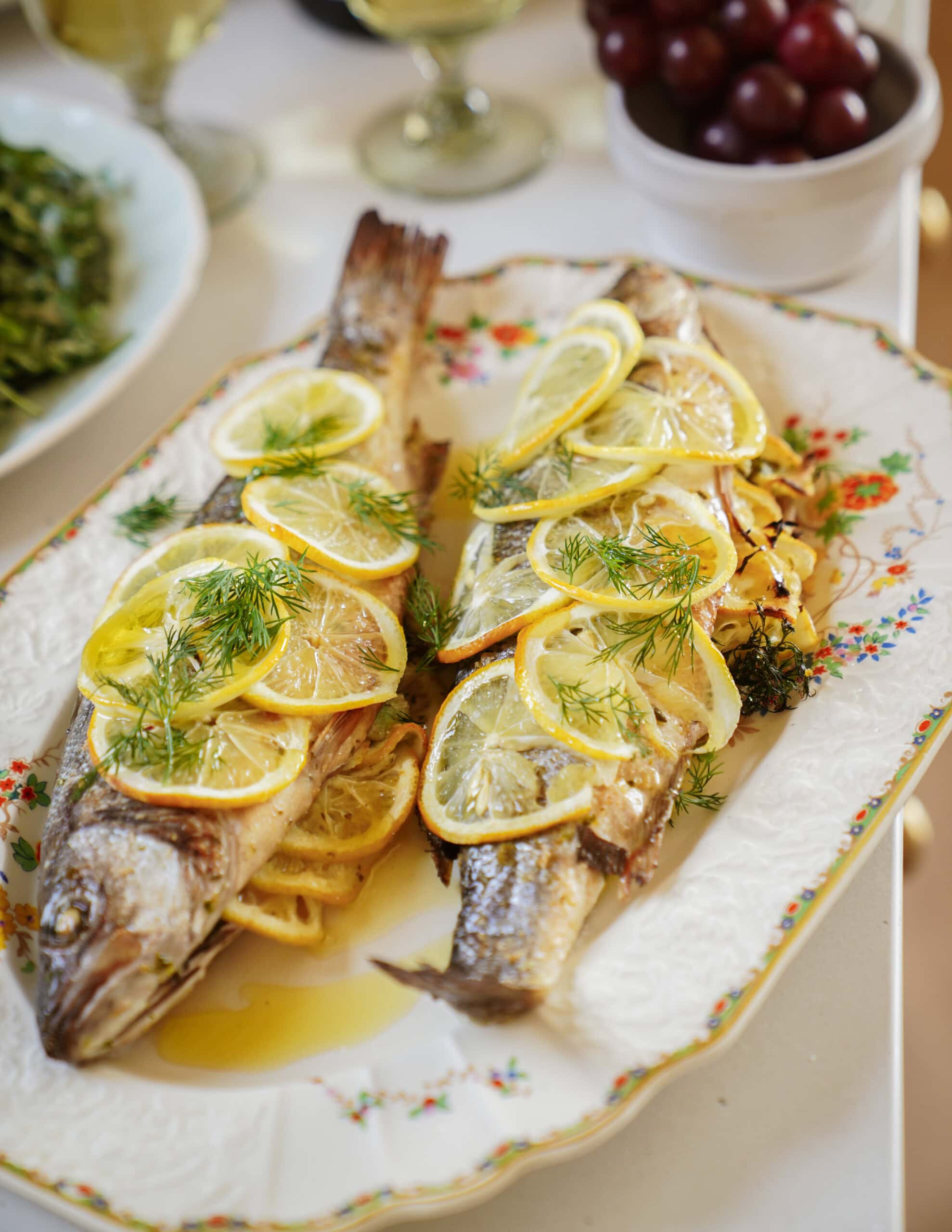 Two whole baked fish garnished with lemon slices, dill, and herbs are served on a floral platter. In the background, there are bowls of salad and red grapes, along with glasses of white wine.