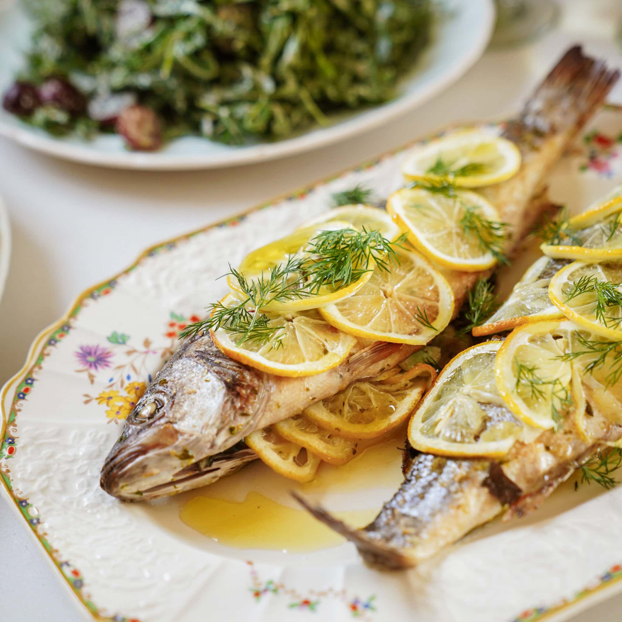 A close-up of two whole baked fish garnished with lemon slices and fresh dill, served on a decorative floral plate. A leafy green salad is visible in the background.