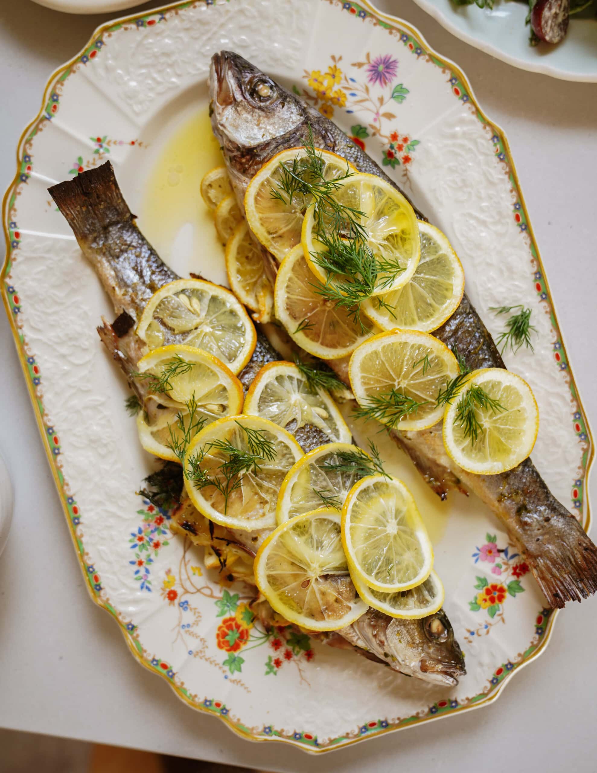 Two whole baked fish garnished with overlapping lemon slices and fresh dill are served on an ornate floral platter. The fish are arranged side by side, surrounded by a patterned border.