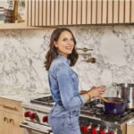 Alex Snodgrass in a denim outfit stands at a stove, smiling at the camera while stirring food in a blue pot. The kitchen has a marble backsplash and beige cabinets.
