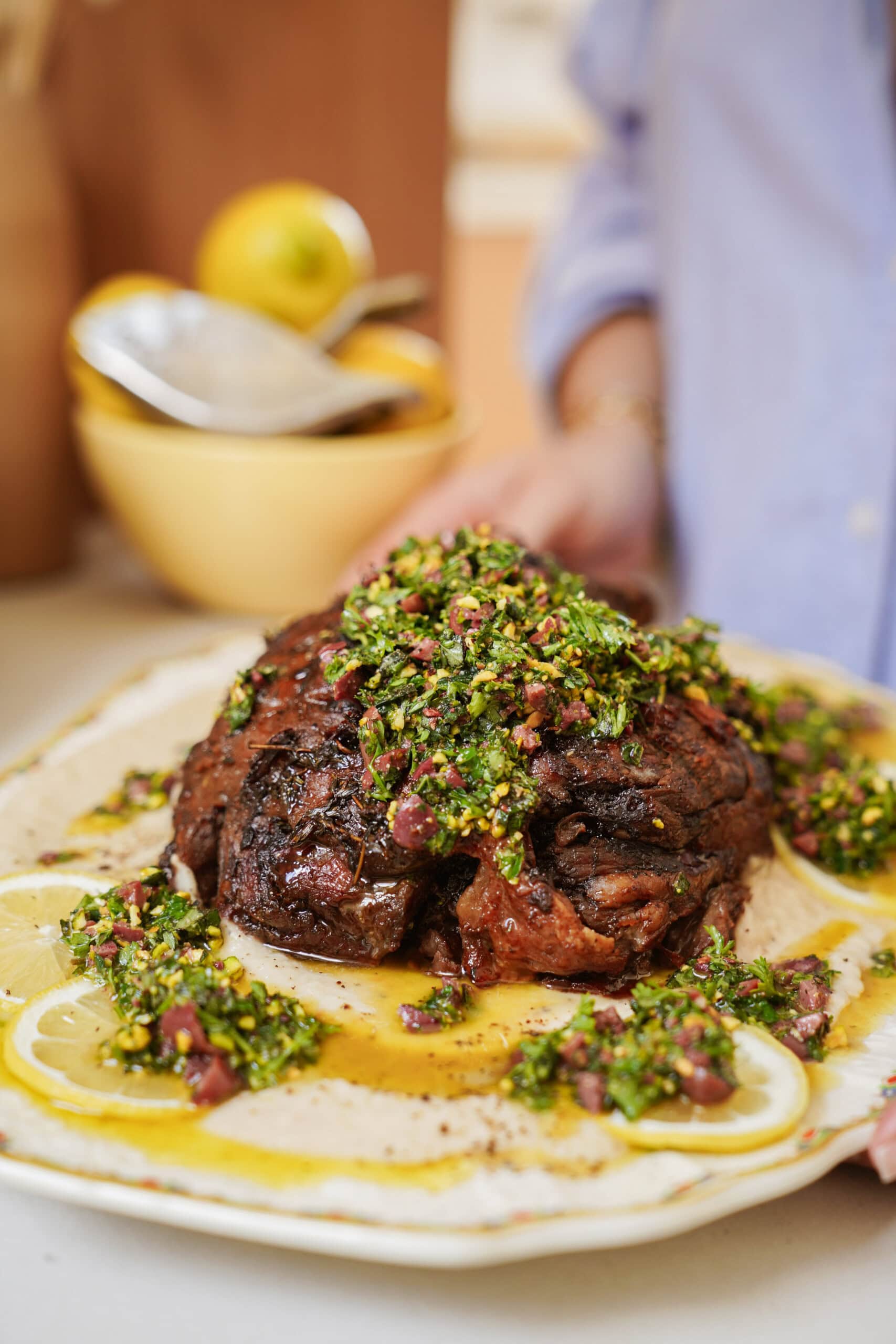 A close-up of a cooked roast topped with a vibrant green herb sauce, served on a plate with sliced lemons, with a person in a blue shirt in the background.