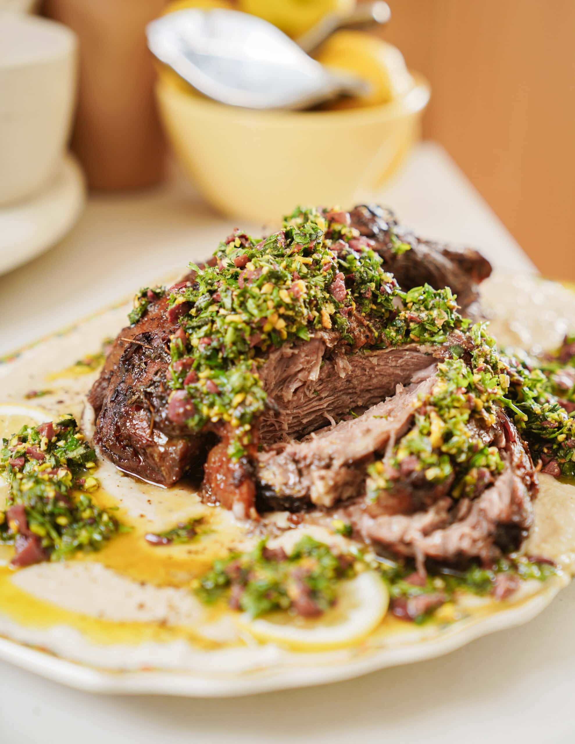 A plate of sliced roast beef topped with a vibrant green chimichurri sauce, served on a bed of creamy sauce with herbs and oil, placed on a table with blurred dishes in the background.