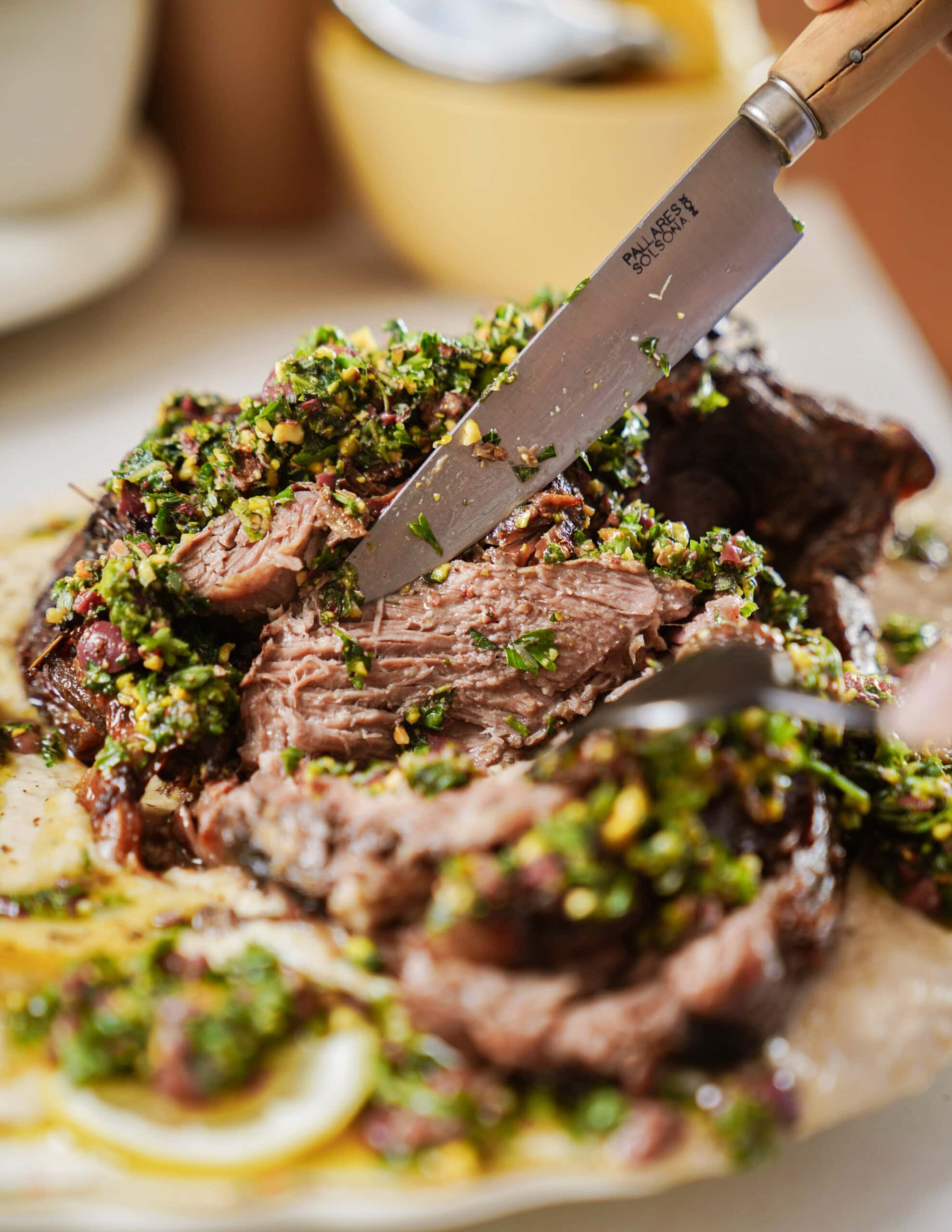 A hand slices into a roast beef topped with a vibrant green herb and nut chimichurri sauce, served on a plate with some visible lemon slices.