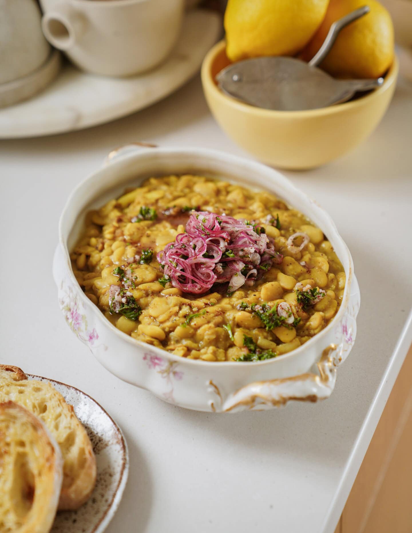 A ceramic dish filled with a chunky yellow stew, topped with pickled red onions and herbs, sits on a kitchen counter near a bowl of lemons and a plate with slices of toasted bread.