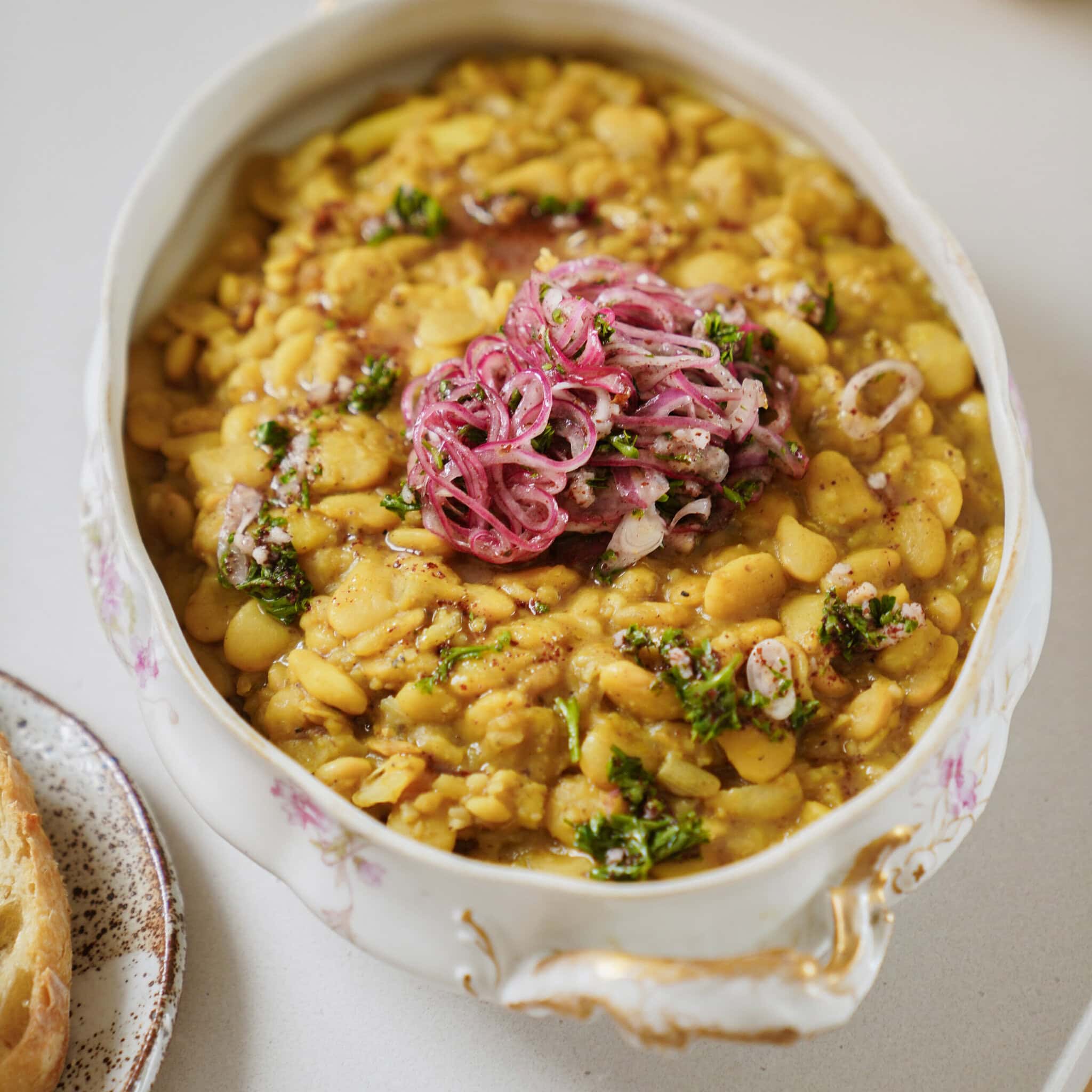 A ceramic dish filled with a yellow bean stew, garnished with pickled red onions and chopped herbs, sits on a light surface. A slice of bread is visible on a plate beside the dish.