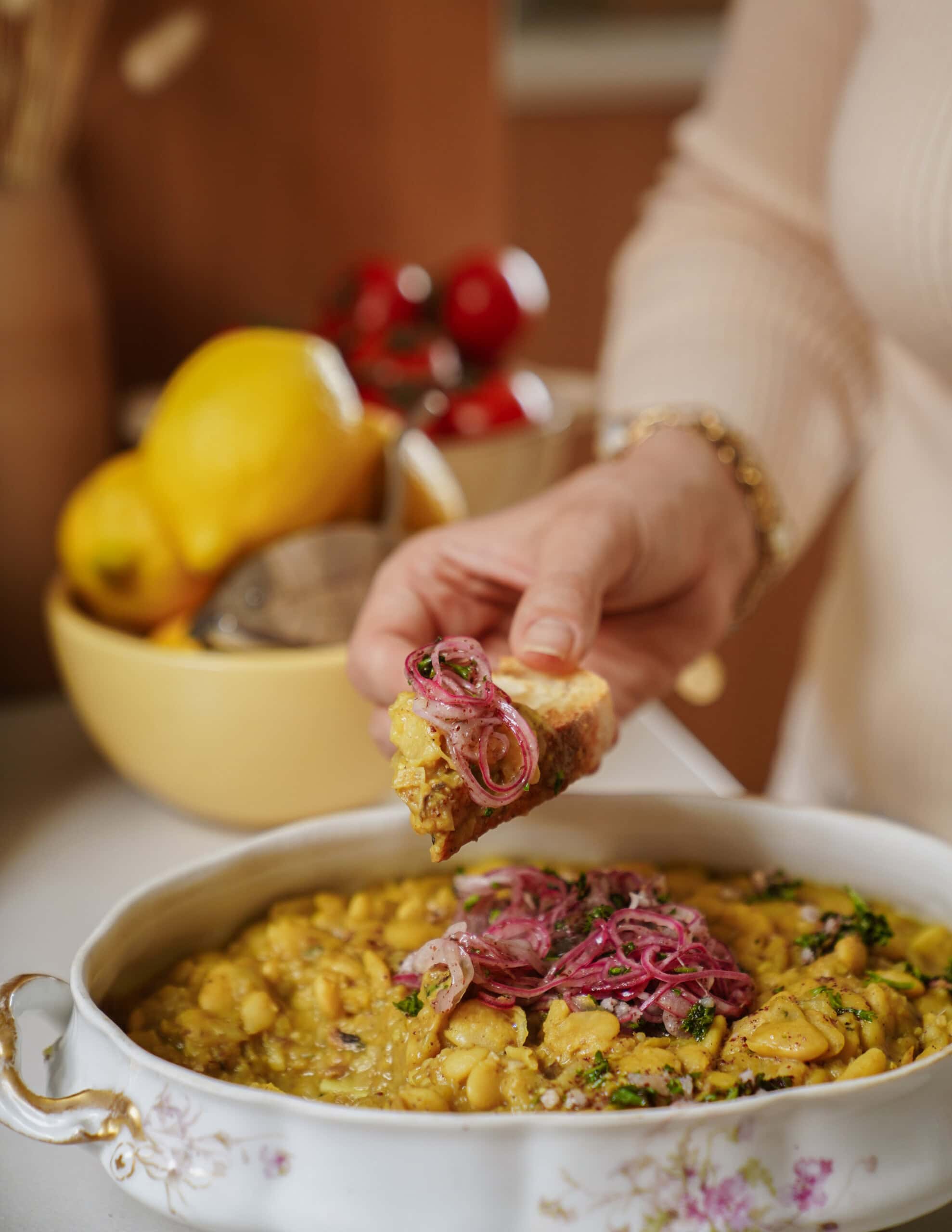 A person holds a piece of bread topped with a yellow bean dish and pickled onions above a serving dish. In the background, lemons and tomatoes are visible in a bowl.