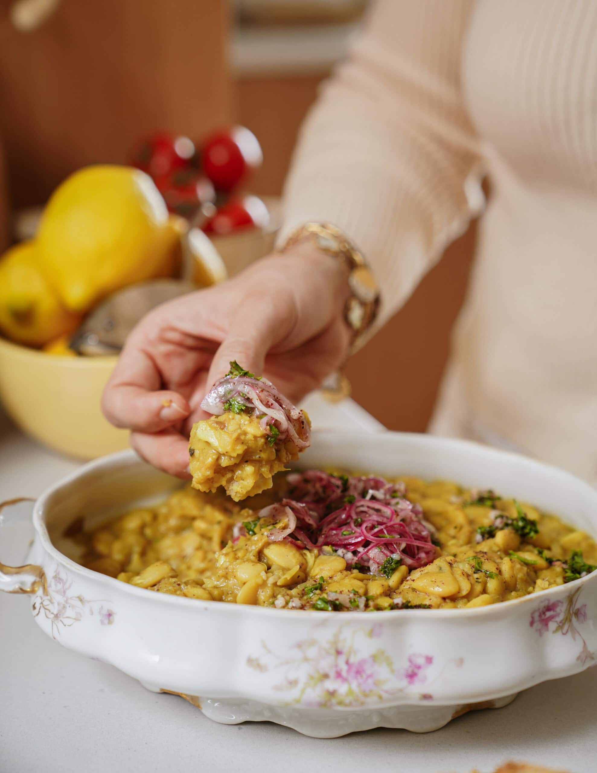 A person scoops a portion of a yellow, mashed dish garnished with pickled onions and herbs from a decorative bowl. Lemons and tomatoes are visible in the background on a kitchen counter.