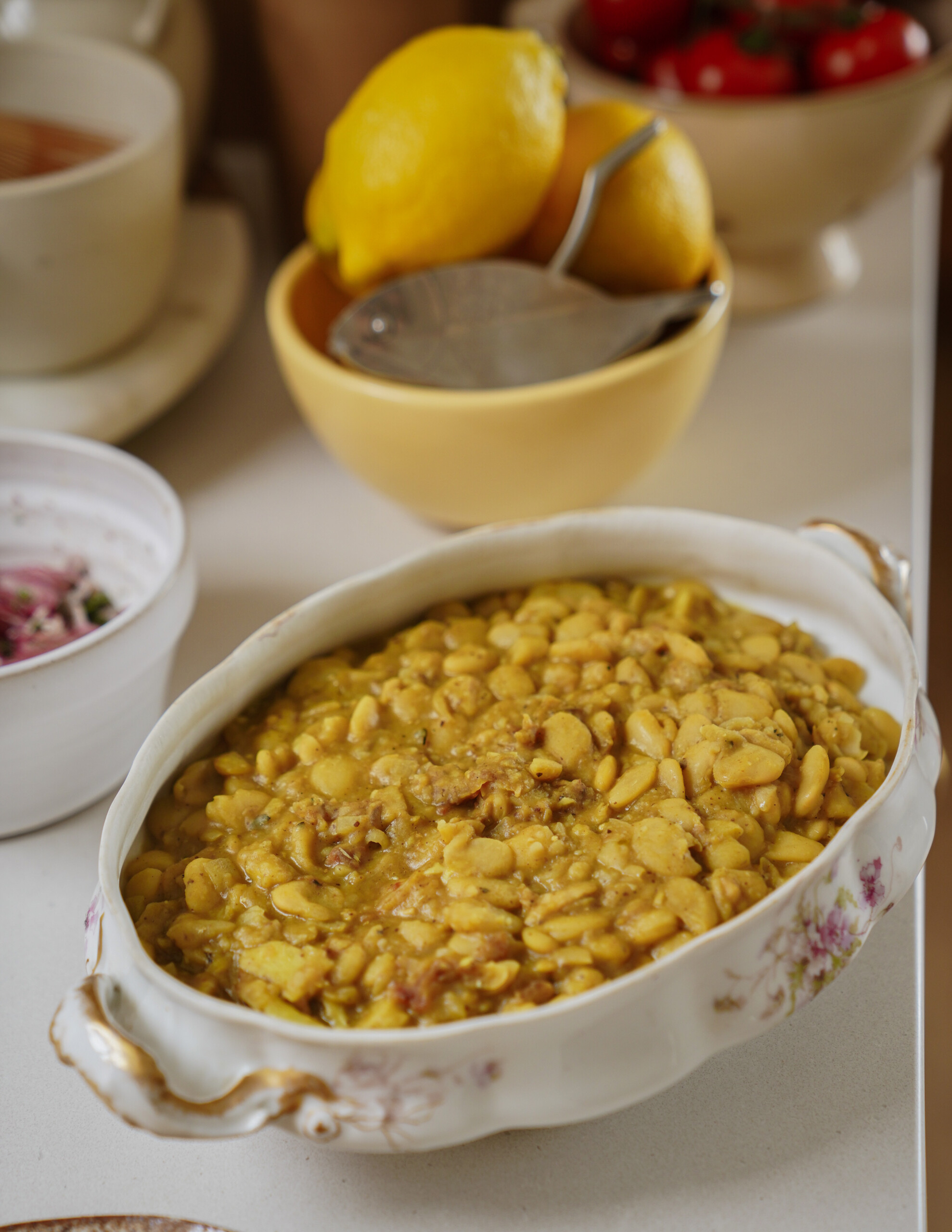 A decorative oval dish filled with a yellow, creamy bean stew sits on a counter. Behind it, a yellow bowl holds whole lemons and a metal citrus squeezer. Other dishes and tomatoes are partially visible in the background.