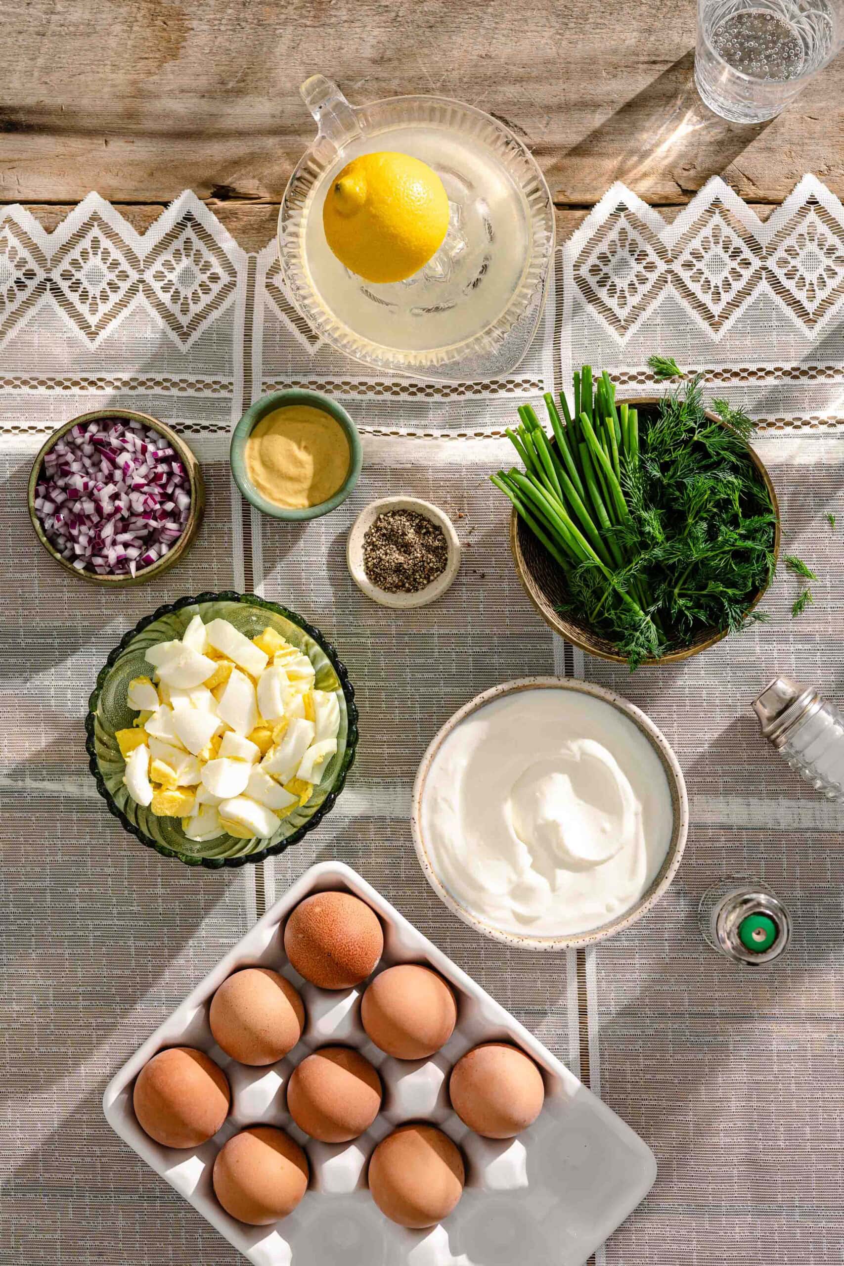 A table set with eggs, chopped red onion, fresh herbs, black pepper, salt, mustard, sour cream, lemon water, and cubed butter on a lace-trimmed tablecloth in bright natural light.