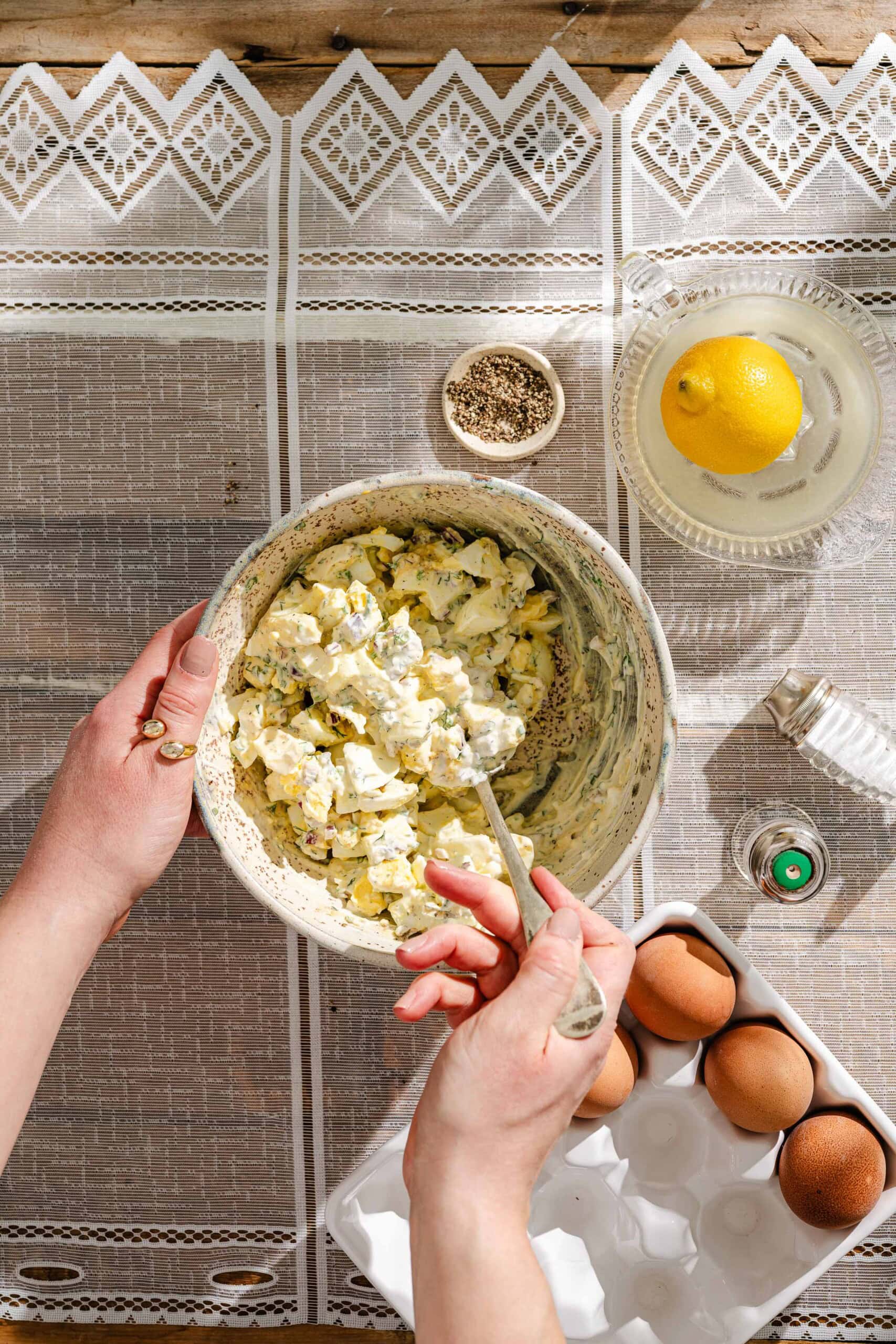 A person mixes an egg salad in a bowl on a patterned tablecloth. Nearby are a lemon, a small bowl of pepper, brown eggs in a carton, a glass bottle, and a juicer.