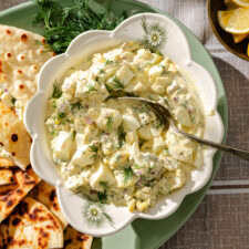 A bowl of creamy egg salad garnished with fresh dill, served with pieces of flatbread on a green plate. Lemon wedges and sprigs of dill are also visible nearby.
