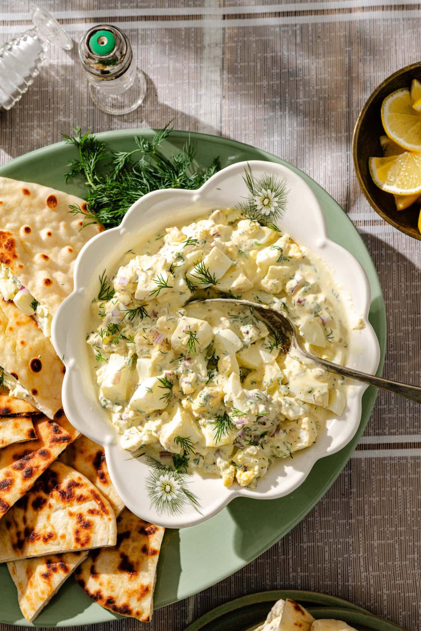 A bowl of creamy egg salad garnished with fresh dill is served with grilled flatbread pieces on a green plate. Lemon wedges and a pepper shaker are nearby on a patterned tablecloth.