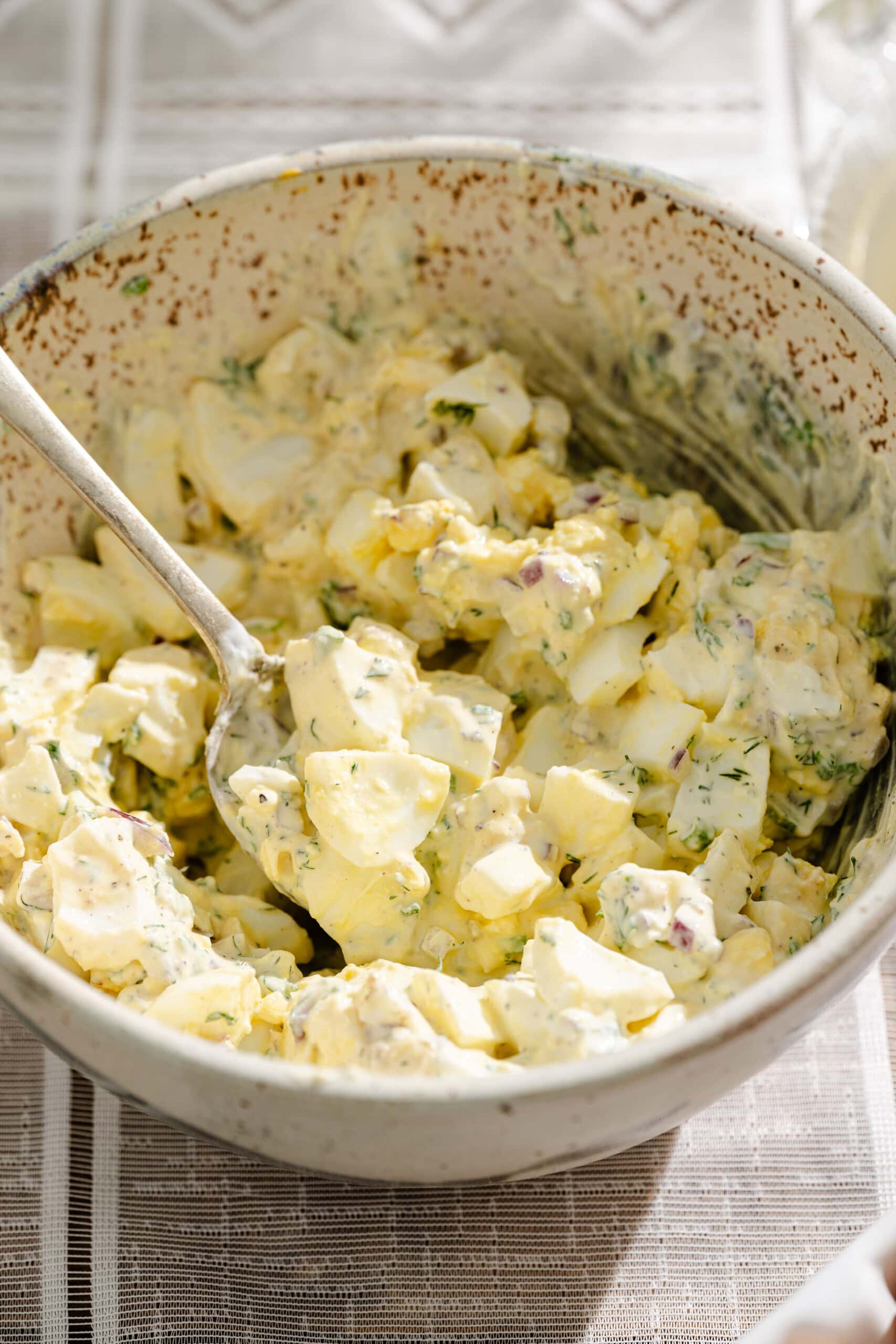 A ceramic bowl filled with creamy potato salad containing chopped potatoes, eggs, herbs, and mayonnaise, with a spoon resting inside the bowl on a patterned tablecloth.