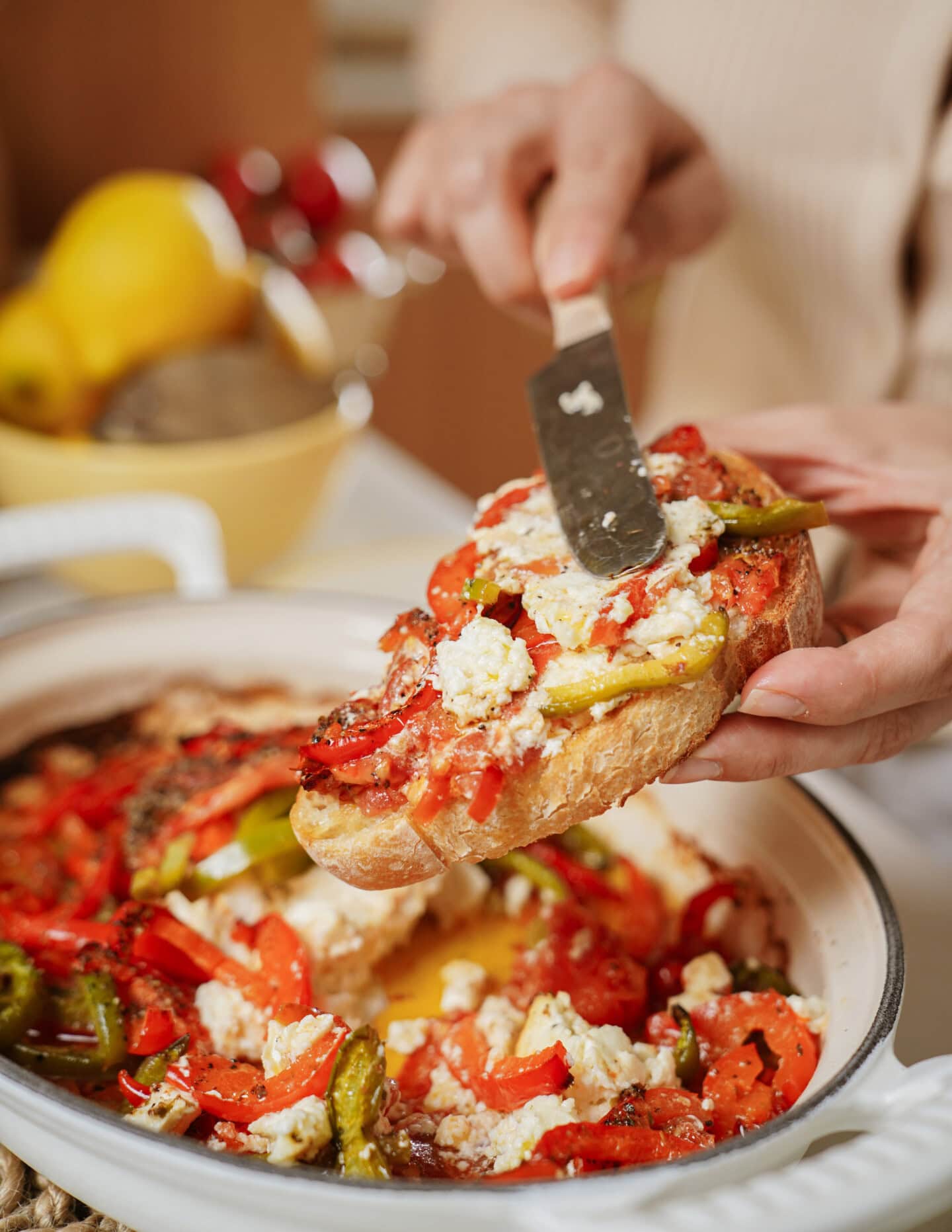 A person spreads ricotta cheese and roasted vegetables on a slice of bread, holding it over a dish filled with more cheese, tomatoes, and bell peppers.