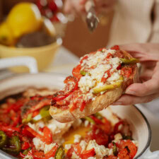 A hand holds a slice of bread topped with baked vegetables and crumbled cheese, with a baking dish of similar food and a lemon squeezer in the background.
