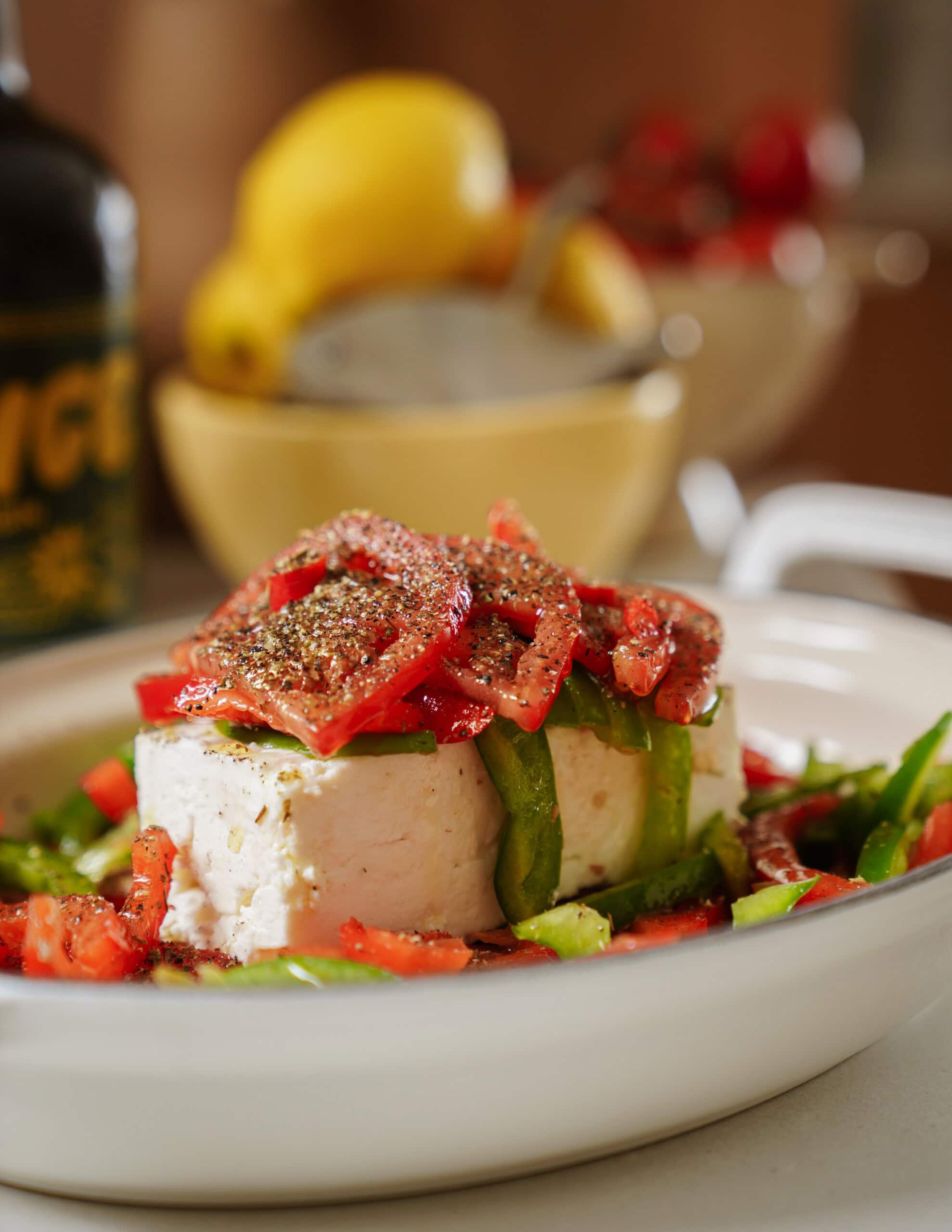 A block of feta cheese topped with sliced red bell peppers and black pepper sits on a white plate, surrounded by green peppers. A lemon, bowl, and bottle are blurred in the background.