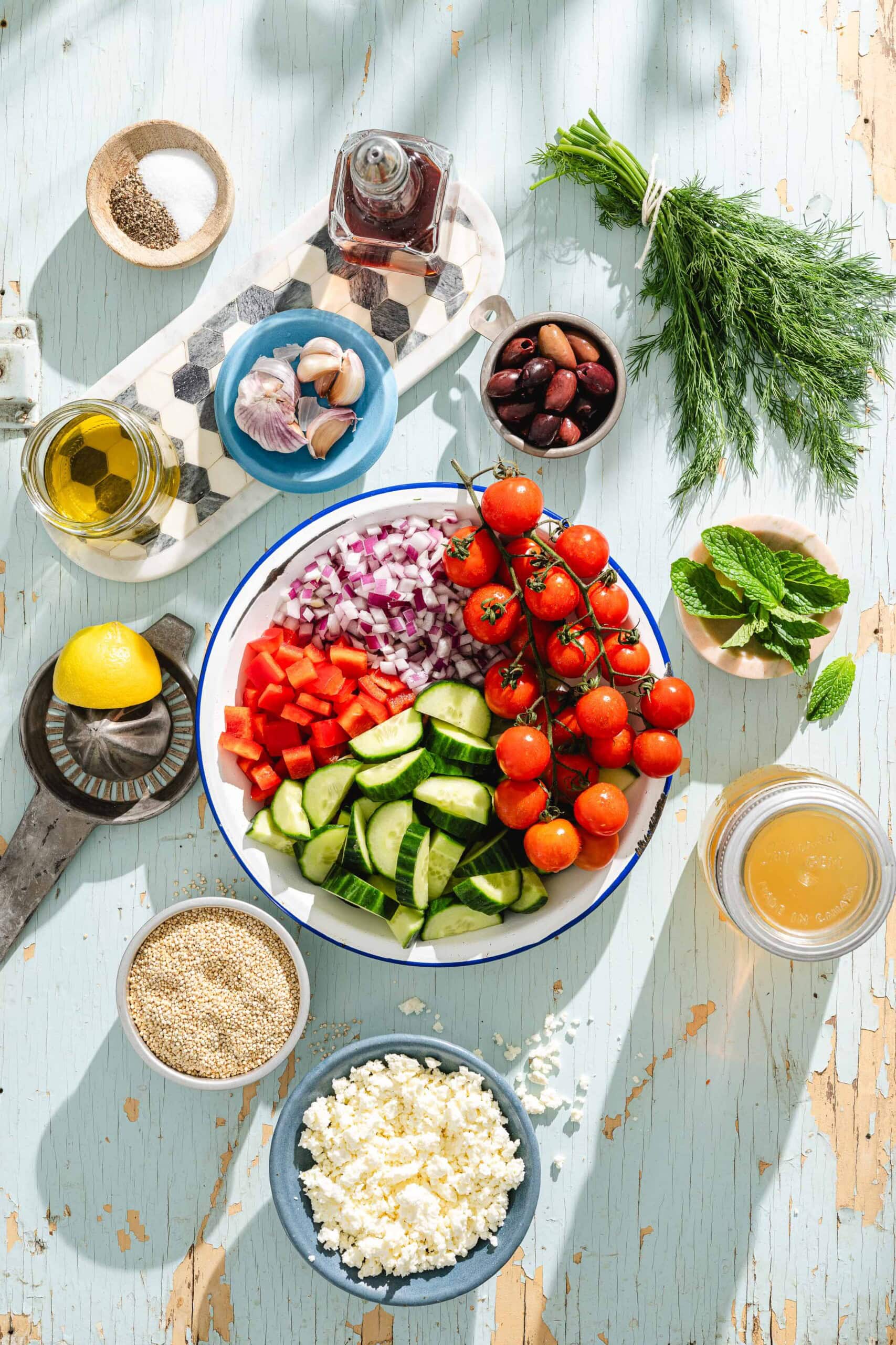 A bowl of chopped cucumber, red pepper, tomato, and red onion sits surrounded by ingredients like olive oil, lemon, feta cheese, olives, quinoa, herbs, garlic, salt, and pepper on a rustic blue table.