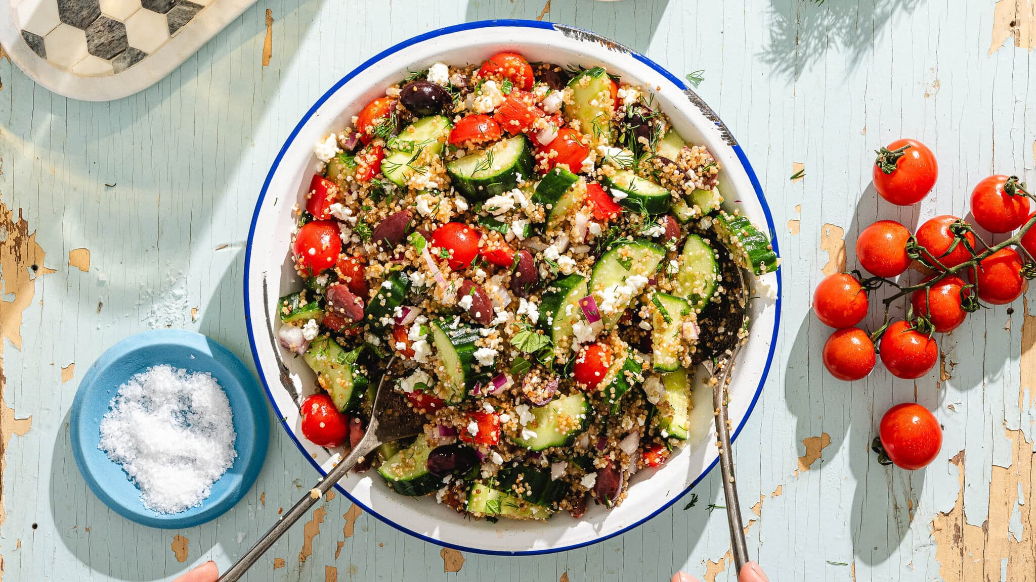 A bowl of Greek salad with cucumbers, tomatoes, olives, quinoa, and feta cheese sits on a rustic light blue table, with a small bowl of salt and a cluster of fresh tomatoes nearby.