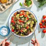 A bowl of quinoa salad with cherry tomatoes, cucumbers, olives, red onions, and feta is being served with utensils. Fresh herbs, salt, and tomatoes are nearby on a light blue wooden table.