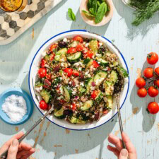 A bowl of quinoa salad with cherry tomatoes, cucumbers, olives, red onions, and feta is being served with utensils. Fresh herbs, salt, and tomatoes are nearby on a light blue wooden table.